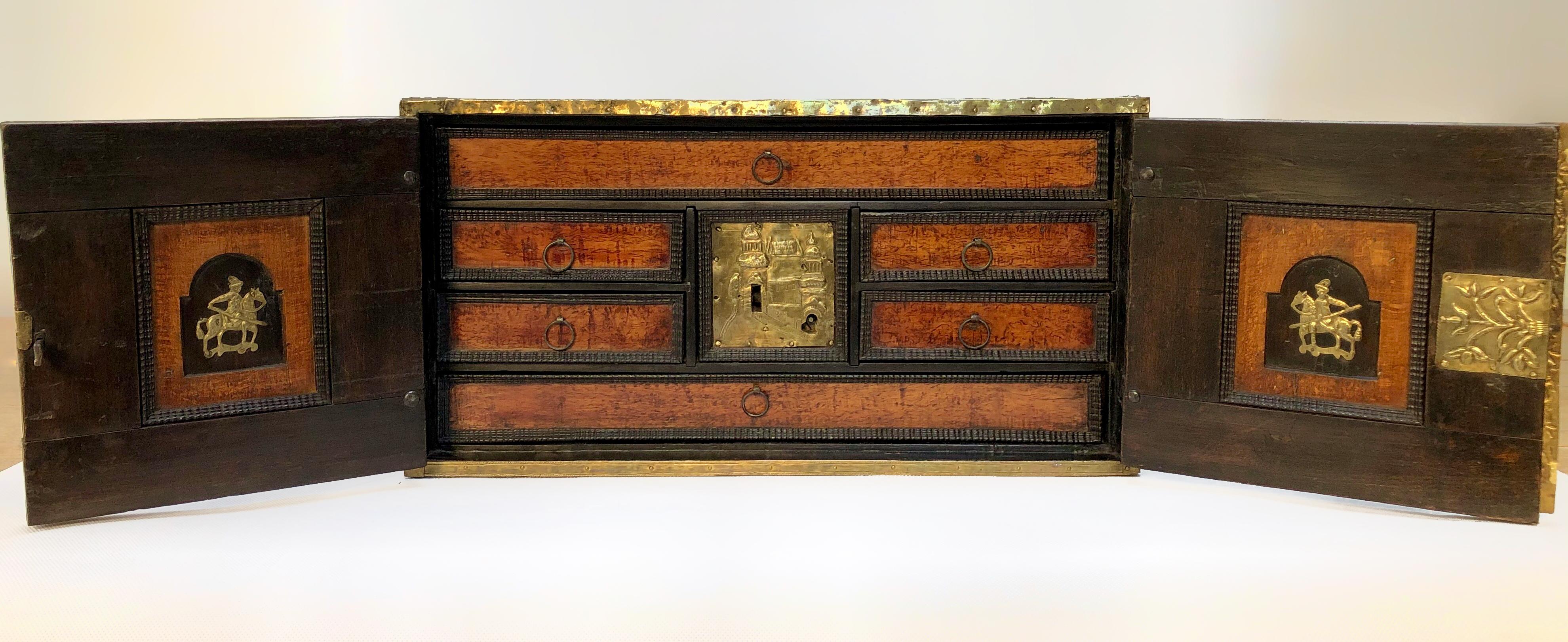 A North European 17th century brass-mounted elm and walnut table cabinet
The pair of doors enclosing a fitted interior of drawers including 'secret' compartments, the central panel decorated with an orientalist brass panel depicting domed