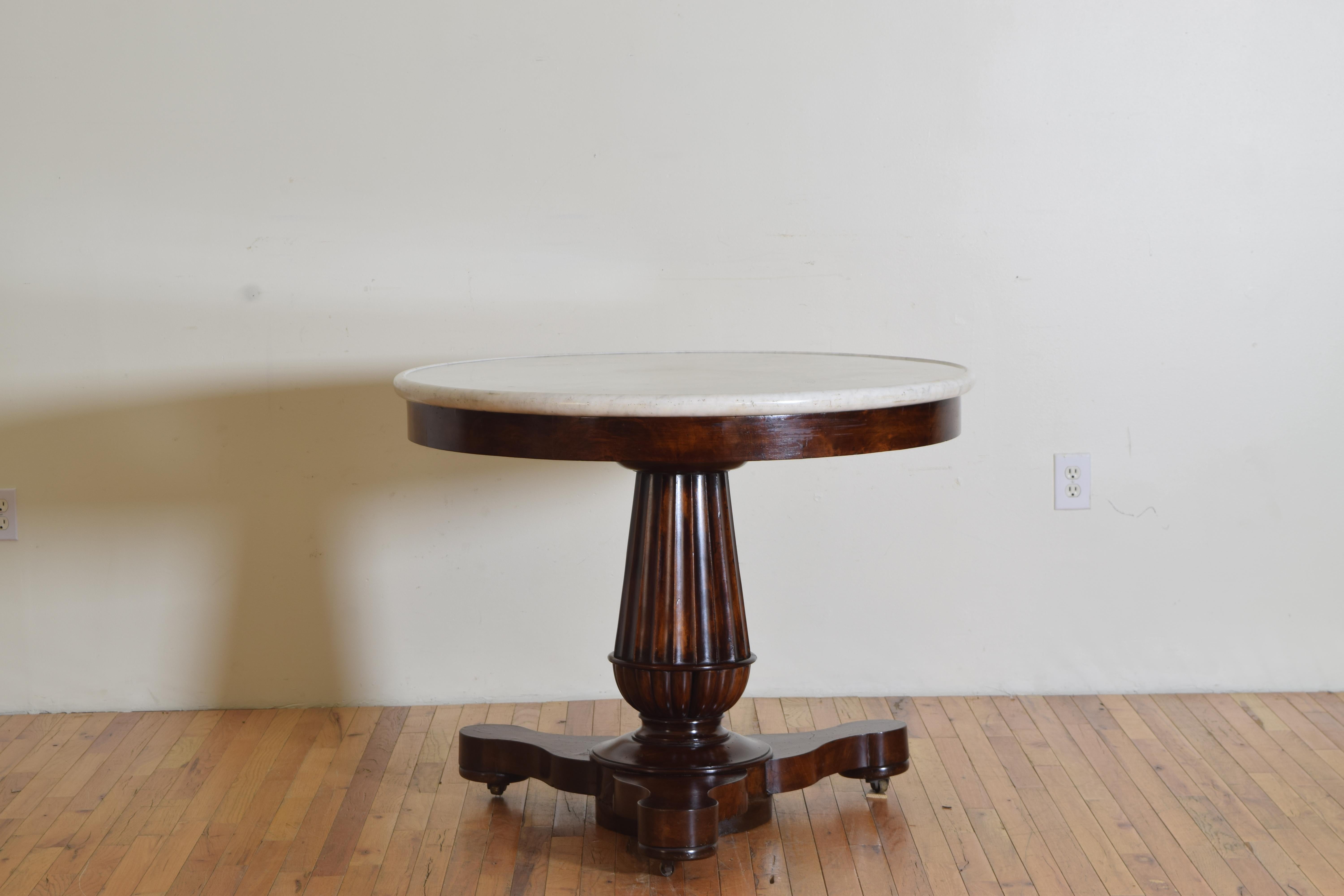 Having a substantial white Carrara marble top with a slightly raised and rounded edge atop a table with a veneered apron and fluted tapering columnar standard resting on a shaped tripartite base with flattened bun feet, raised on original brass