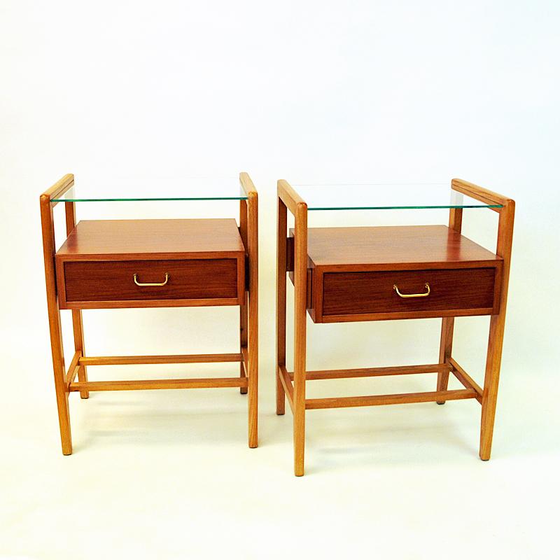 Lovely and classic pair of Norwegian lacquered and stained teak and beech bedside - side table tables from the 1950s. The tables has solid beech legs, shelves with brass handles and a clear glass shelf on top. Magazine/newspaper shelf underneath.