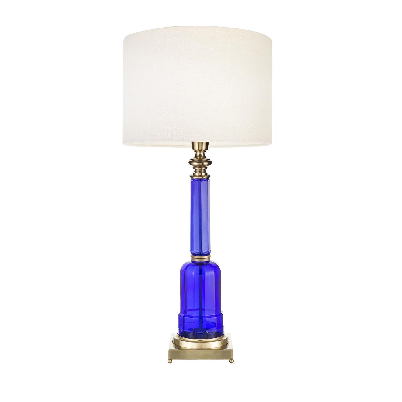 This striking Novecento lamp in a luxurious deep blue is a beautifully decorative item in brass and colored glass. The 17 cm square pedestal with golden ball feet support the glass body of the lamp and the lampshade in clear fabric is covered with