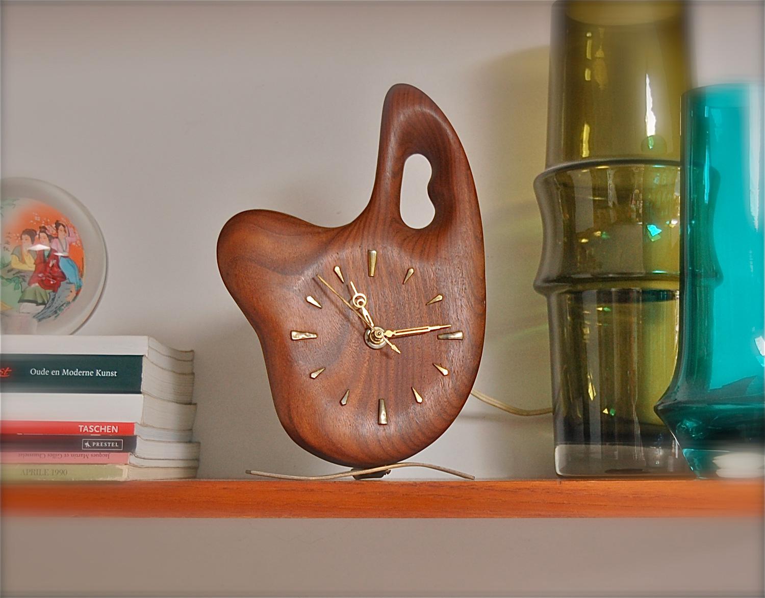 A novelty table or desk clock by German manufacturer Jacob Palmtag. An organically shaped piece of solid timber, resembling an artist palette, on brass feet. The slightly raised strips of brass round the periphery of the face indicate the hours. The