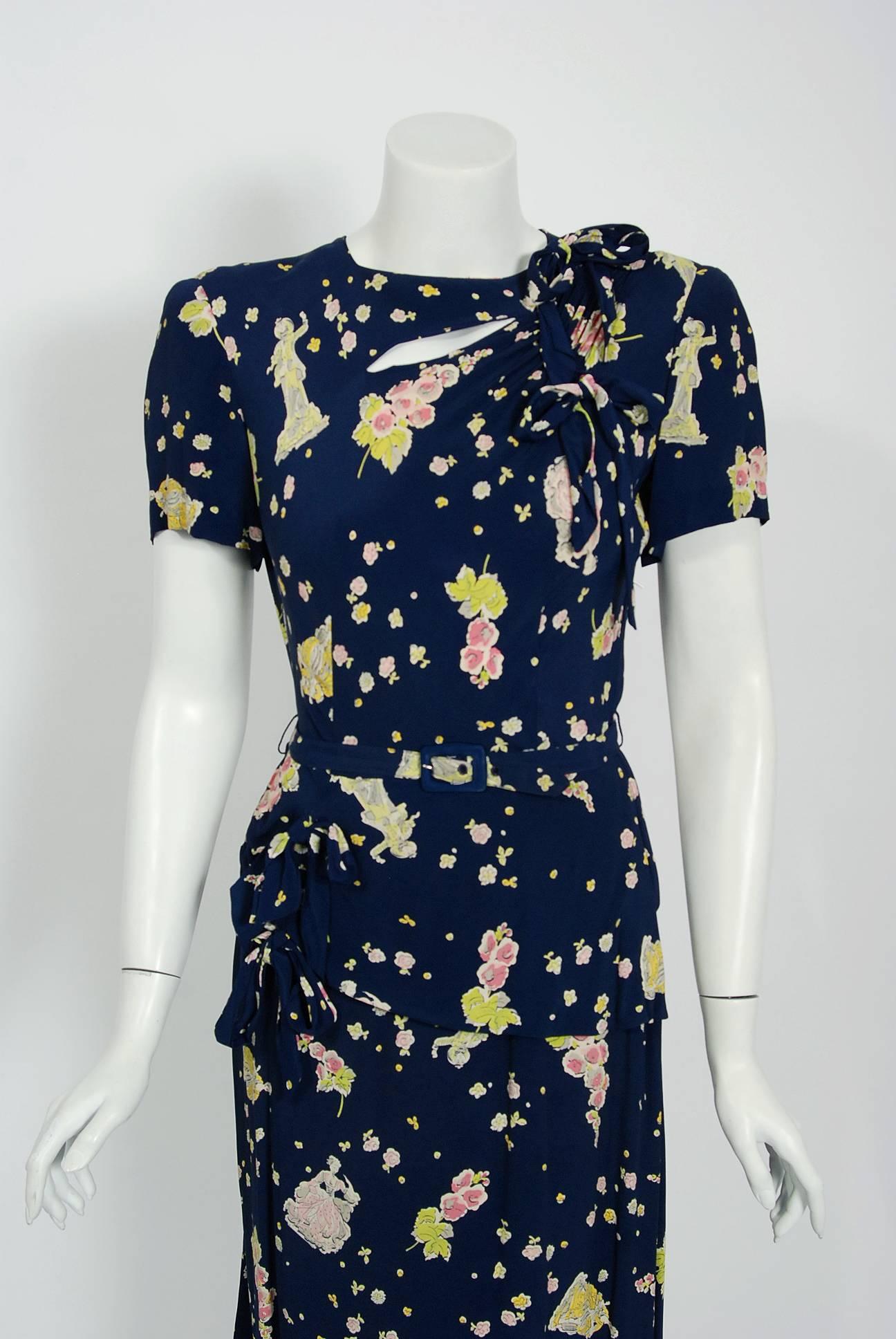 With its adorable Victorian dolls scenic novelty print and flawless styling, this Trudy Hill designer navy-blue rayon dress has the casual elegance the 1940's were known for. The short sleeve asymmetric cut-out bow trimmed bodice is very flattering