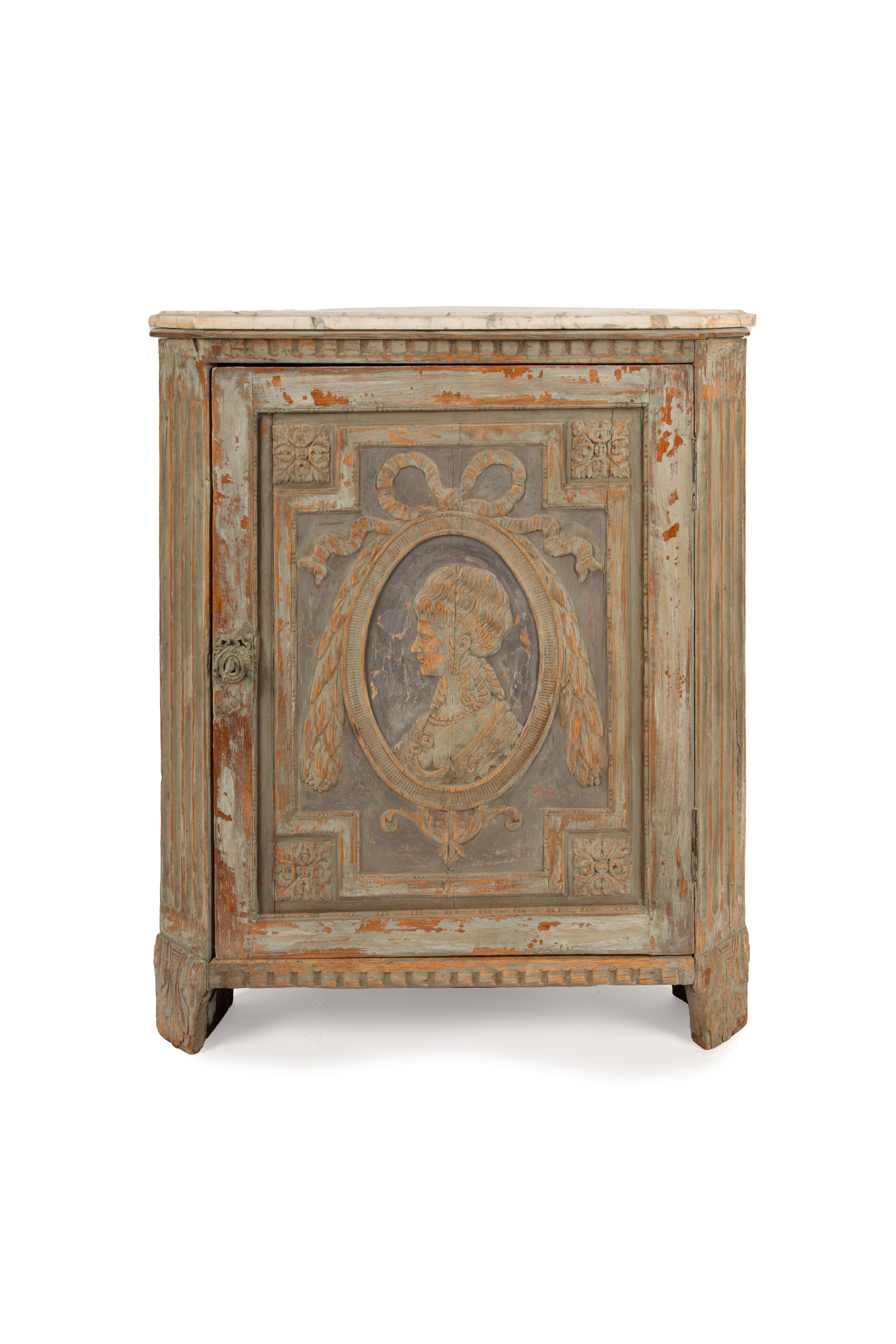 This elegant 18th to early 19th-century Gustavian corner cabinet features exquisite hand-carved details, accentuated by its original patinated finish and a gracefully aged appearance. The central panel showcases a finely carved female profile set