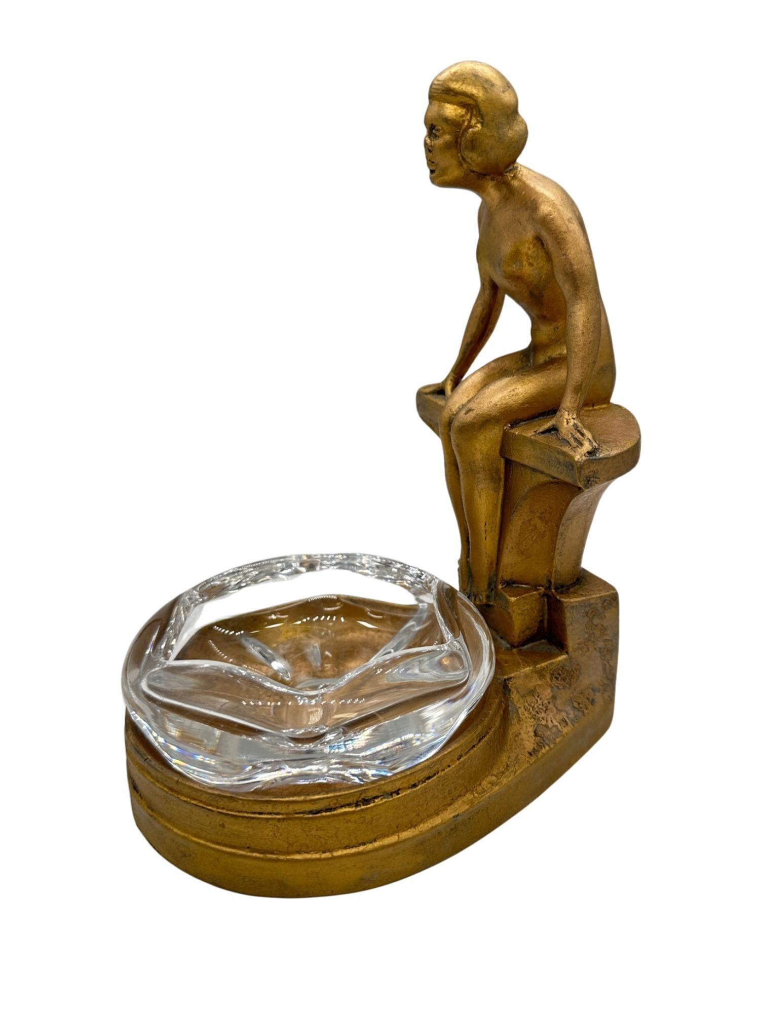 A stunning Art Deco 'NuArt Creations' figural female nude from the 1930s gracefully sits by a pool. This metallic piece, originally designed as an ashtray, features an elegant bronze finish and prominently displays the 'NuArt Creations' emblem at