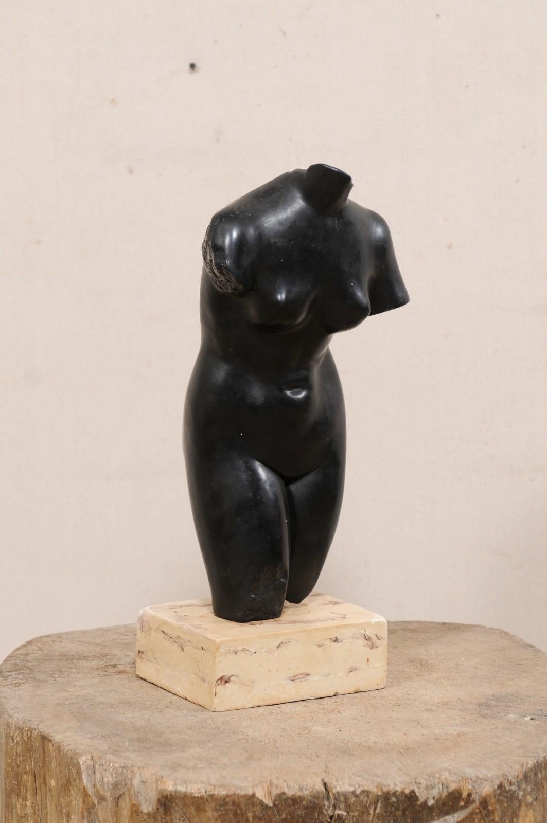 A vintage European nude female torso sculptural art piece. This stone composite statue, which depicts a shapely female torso, or mid region, done in black, and raised upon a contrasting neutral colored squared base. This artisan feminine piece from