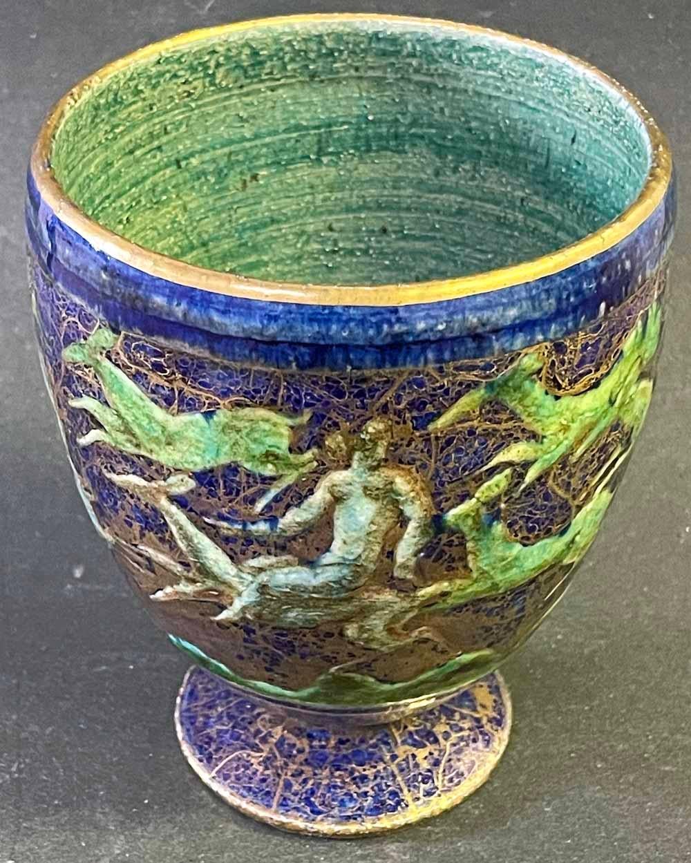 Gorgeously glazed in jewel tones -- sapphire blue, emerald green, and gold -- this footed urn or vase by the great Jean-Leon Mayodon depicts a line of three female nudes riding Art Deco deer, traveling around the surface of the piece and surrounded