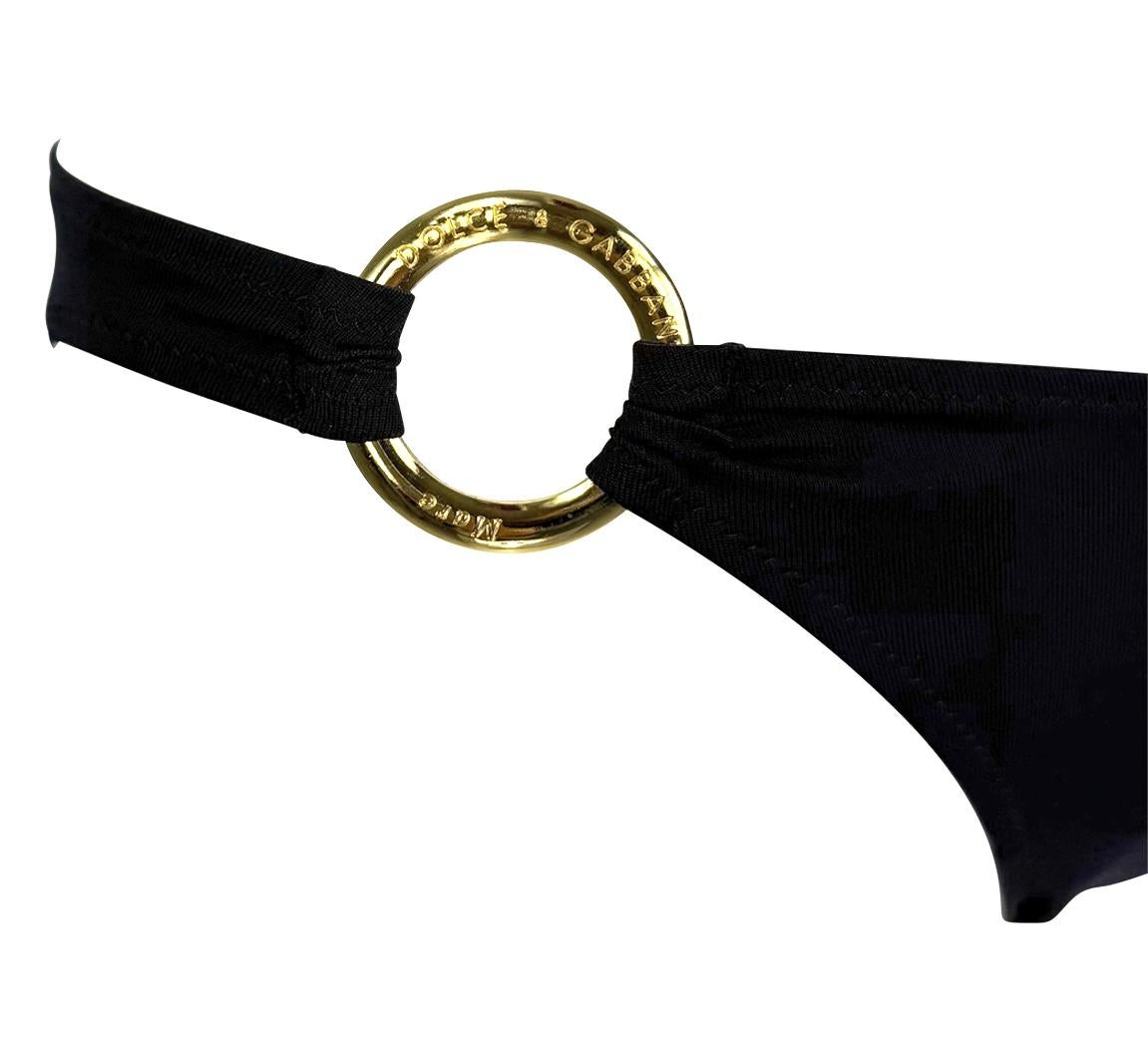 This unworn black stretch bikini set from the early 2000s by Dolce & Gabbana features a classic triangle halter top paired with low-rise bottoms. Gold-tone logo-engraved rings accent each hip, adding a touch of branded glamour to this minimal