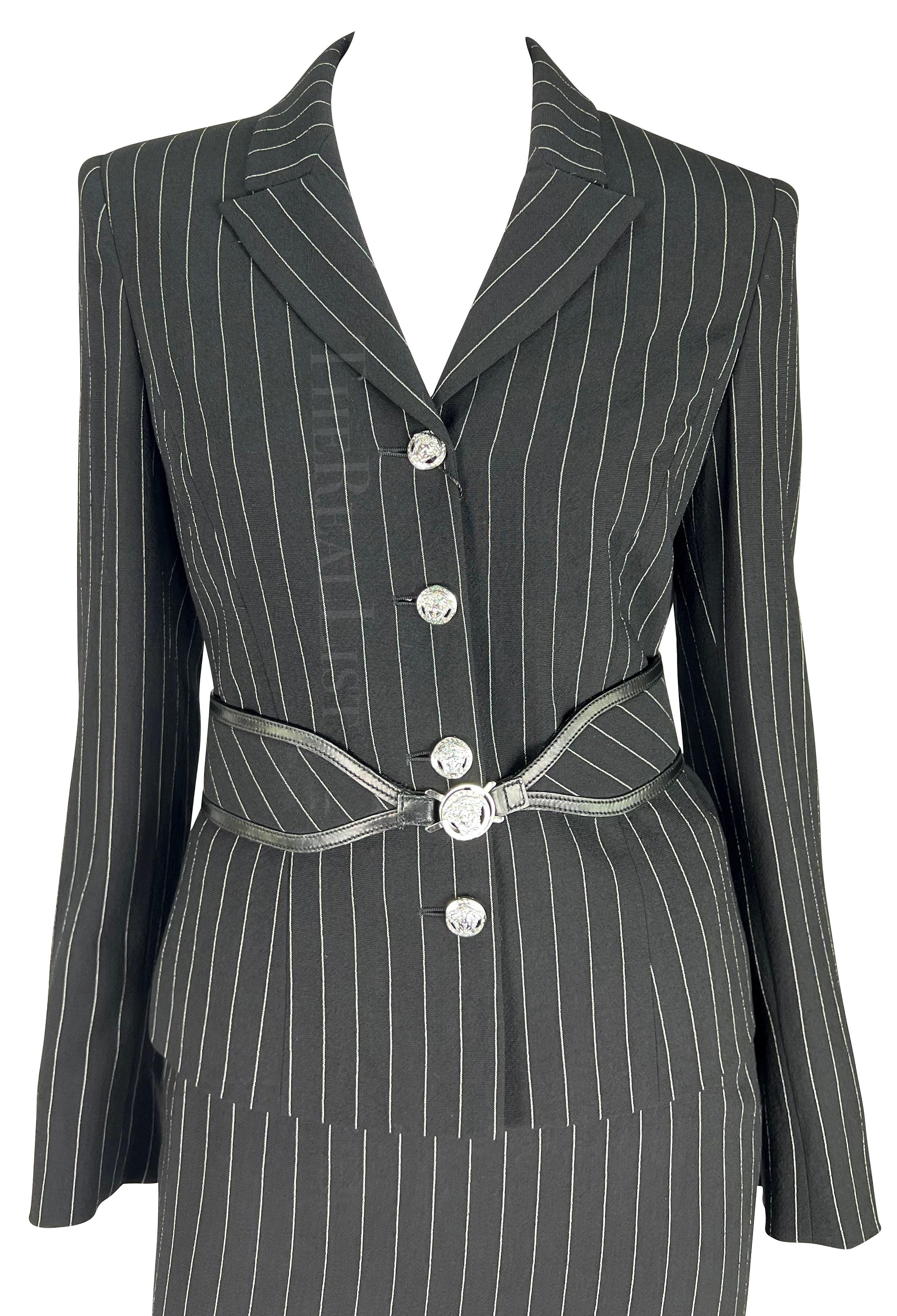 Presenting a black wool pinstripe Versace skirt suit, designed by Donatella Versace. From the Fall/Winter 2004 collection, this coordinated ensemble includes a pinstriped blazer, skirt, and belt. Its traditional silhouette is uplifted by shiny