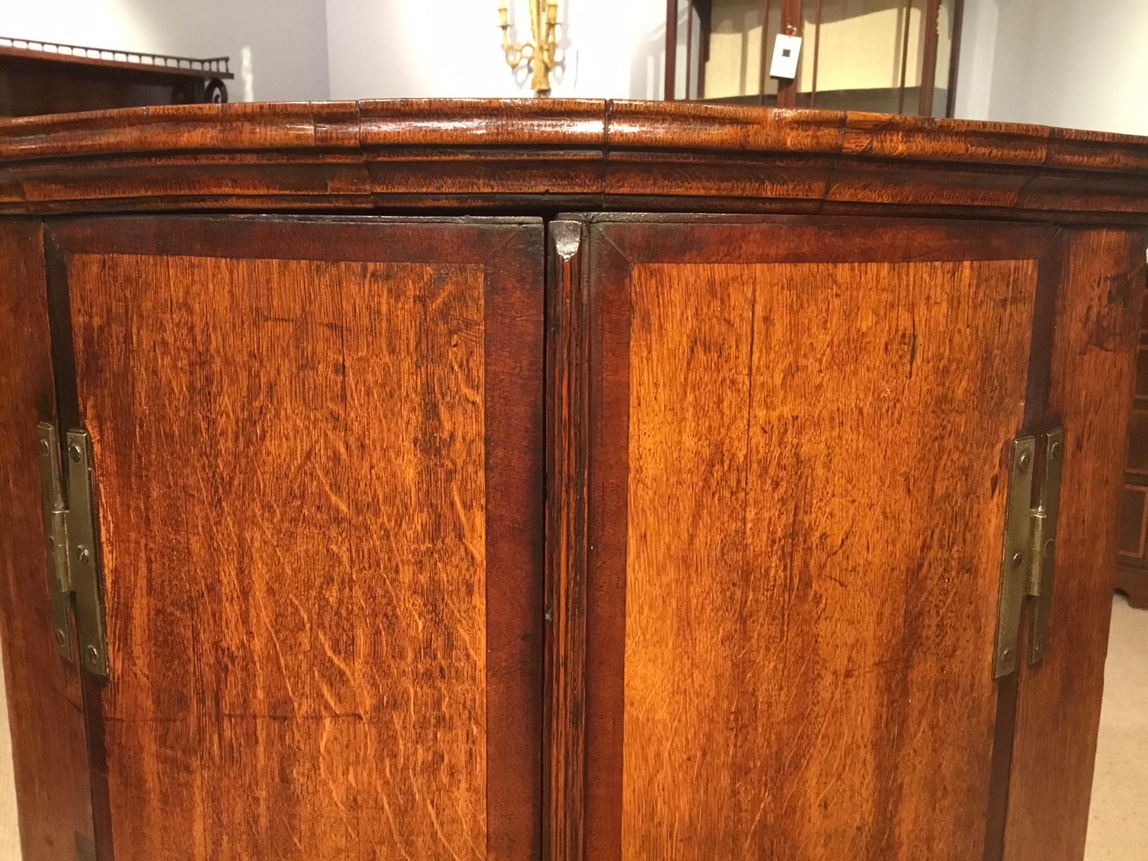 An oak and mahogany cross banded George III Period corner cupboard. A good example of a George III Period bow front corner cupboard with oak doors cross banded in mahogany and retaining the original H hinges, opening to reveal four oak shelves,