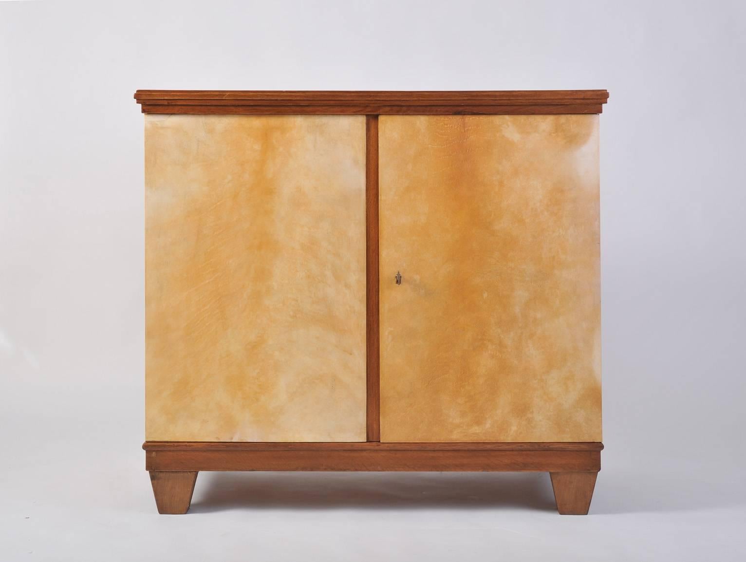 An oak and velum two-door cabinet, in the manner of Jean Michel Frank, the case clad in velum on all sides, topped by a stained oak top with moulded edges, resting on four solid square tapering feet, revealing a central shelf, the doors internally