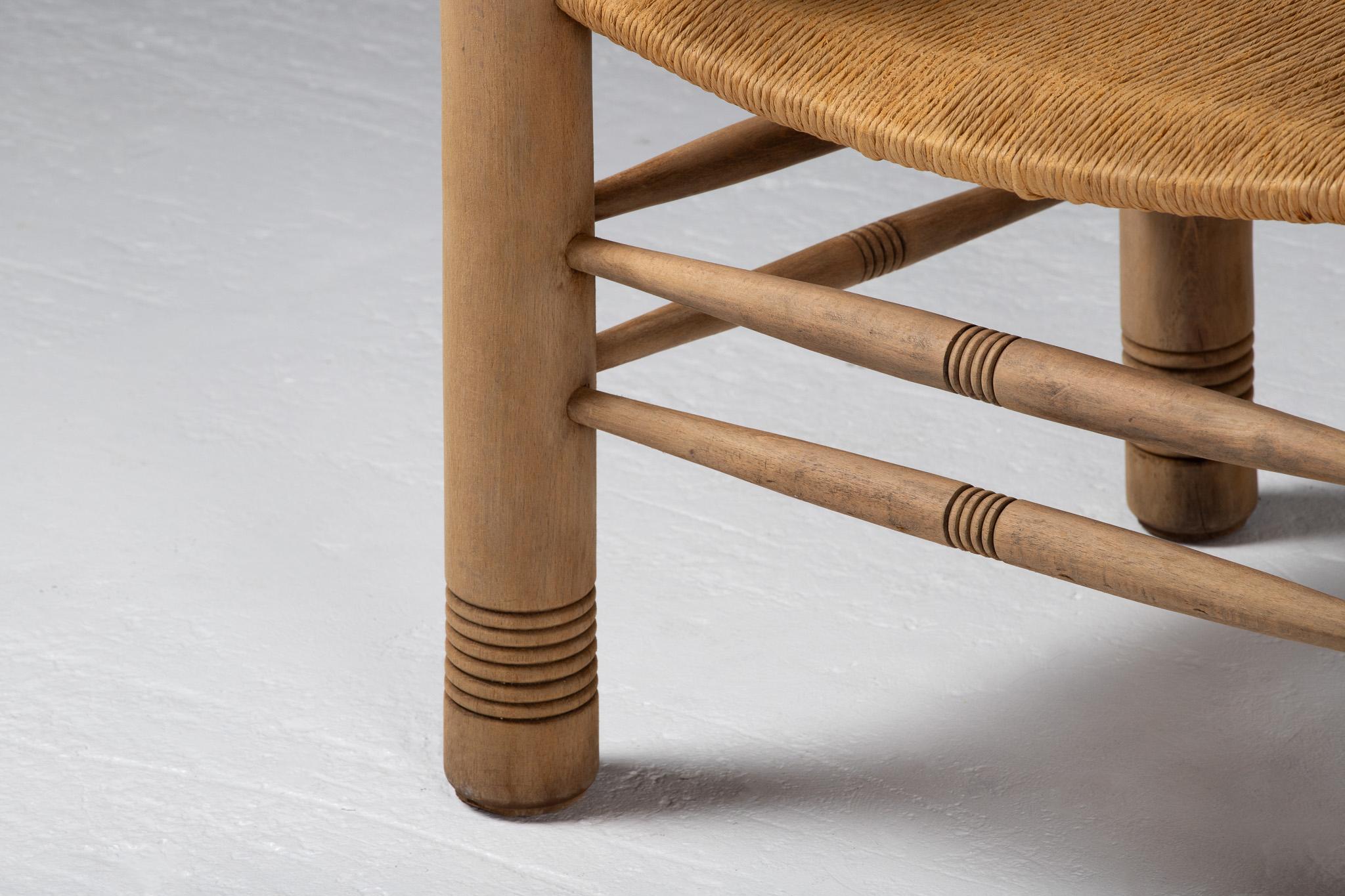 A quiet monument to French rustic modernism, this armchair draws the eye with its deliberate naïveté—rounded finials perched atop robust uprights, sweeping armrests that arc with softened geometry, and legs scored with concentric bands that recall