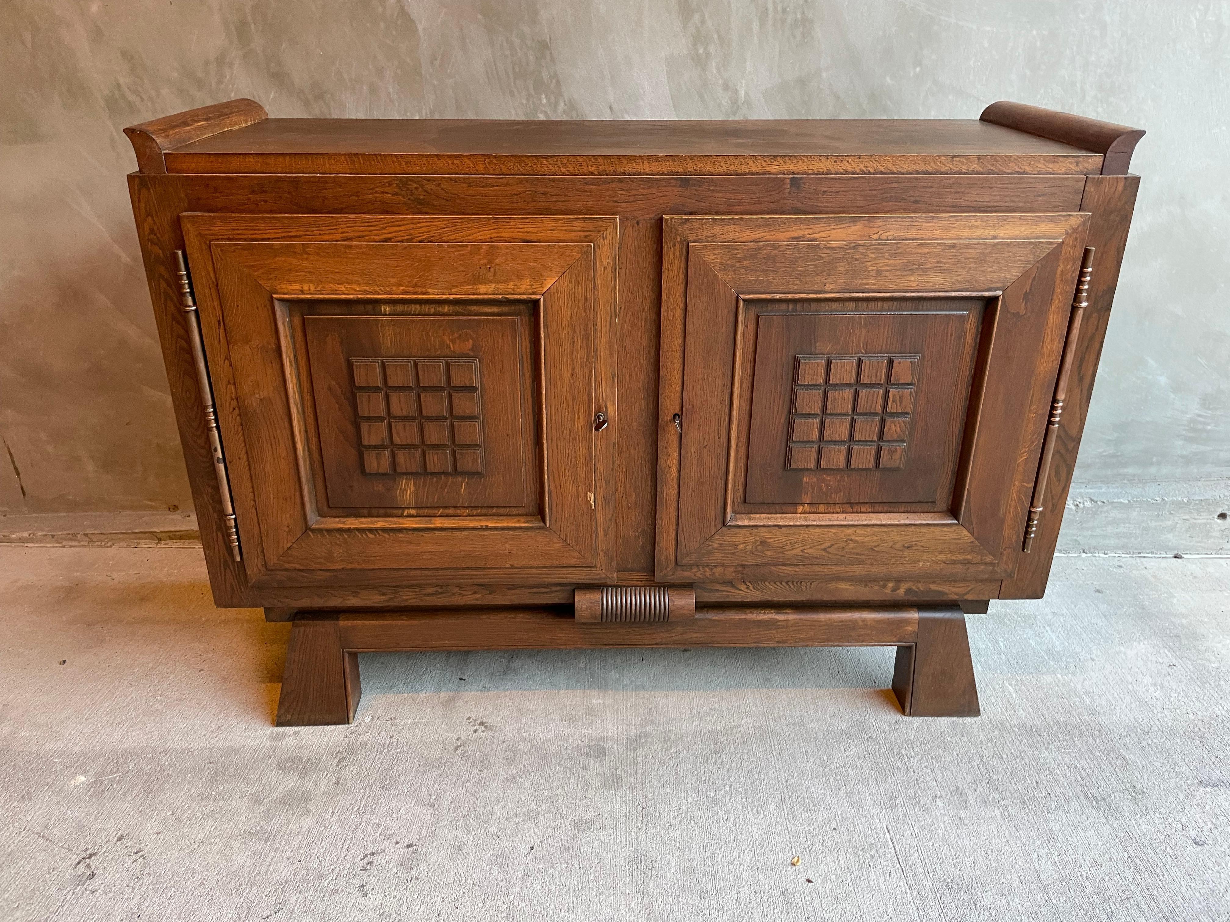 Two door Brutalist Art Deco era oak sideboard by Charles Dudouyt in all original condition. Solid oak construction throughout with lathe turned elongated copper hinges contrasted by geometrical motifs on the frame and panel door fronts. Interior has