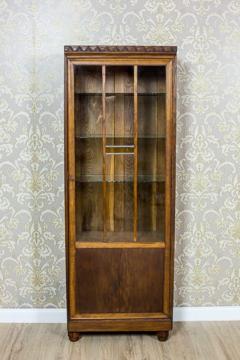 A small, one-door piece of furniture, circa 1930, with a glazed door.
This amazing bookcase is made in oak and oaken burl.
The simple shape of the bookcase has been varied only by a geometrical ornament on the top, and an insert of colored glass