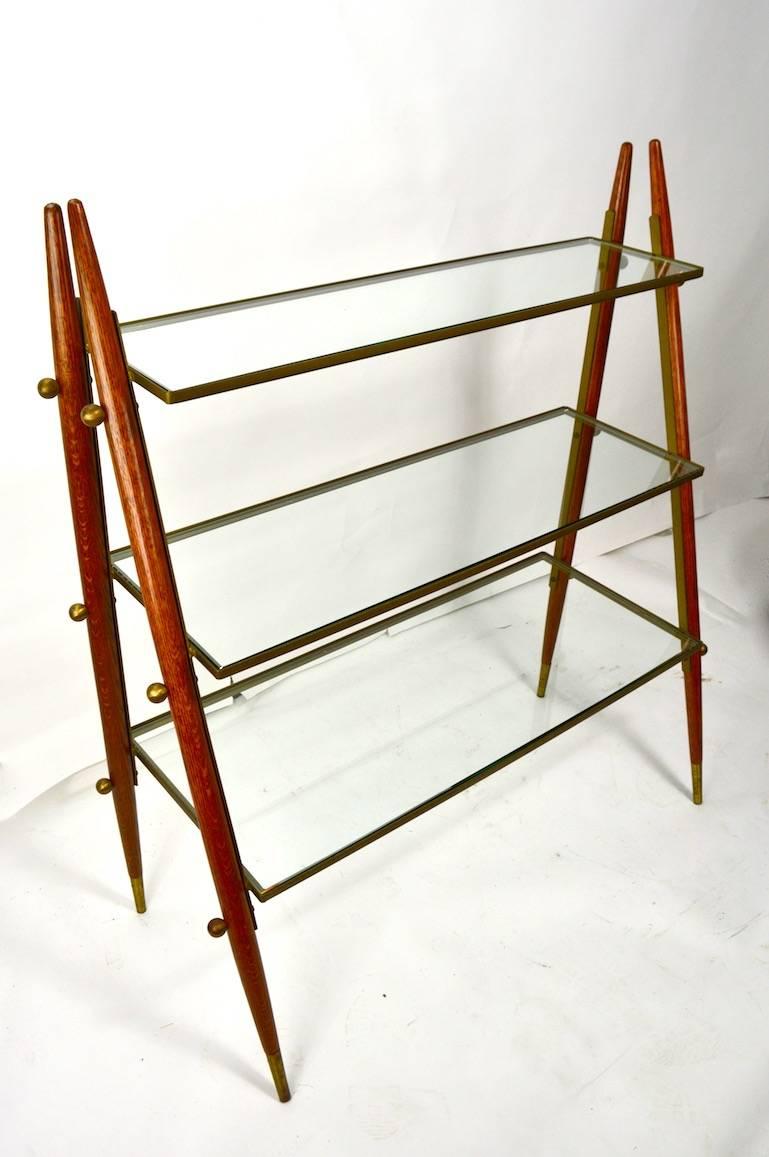 Stylish and chic Mid-Century Modern three level shelf storage, display unit, with oak, steel and brass frame. Each shelf has thick ( 3/8 inch ) plate glass which rests in an angle steel frame, shelves are graduated in size. The tapered pole legs are