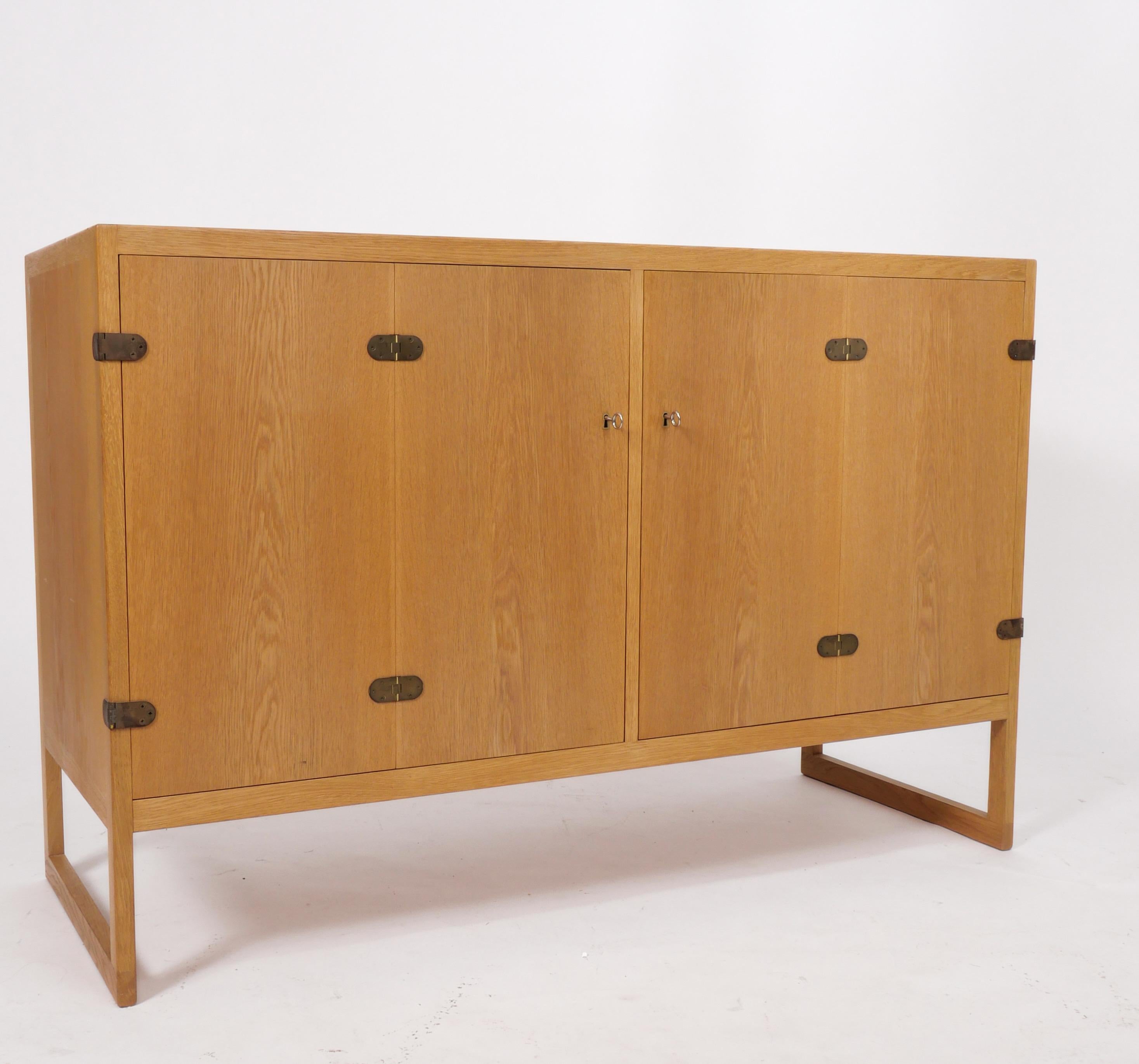 Beautiful cabinet in Oak by Børge Mogensen. Patinated brass hinges at front; locking, folding doors open outward to reveal adjustable shelving as well as two shallow felt-lined drawers. Both door keys present. The cabinet rests on a runner base; the