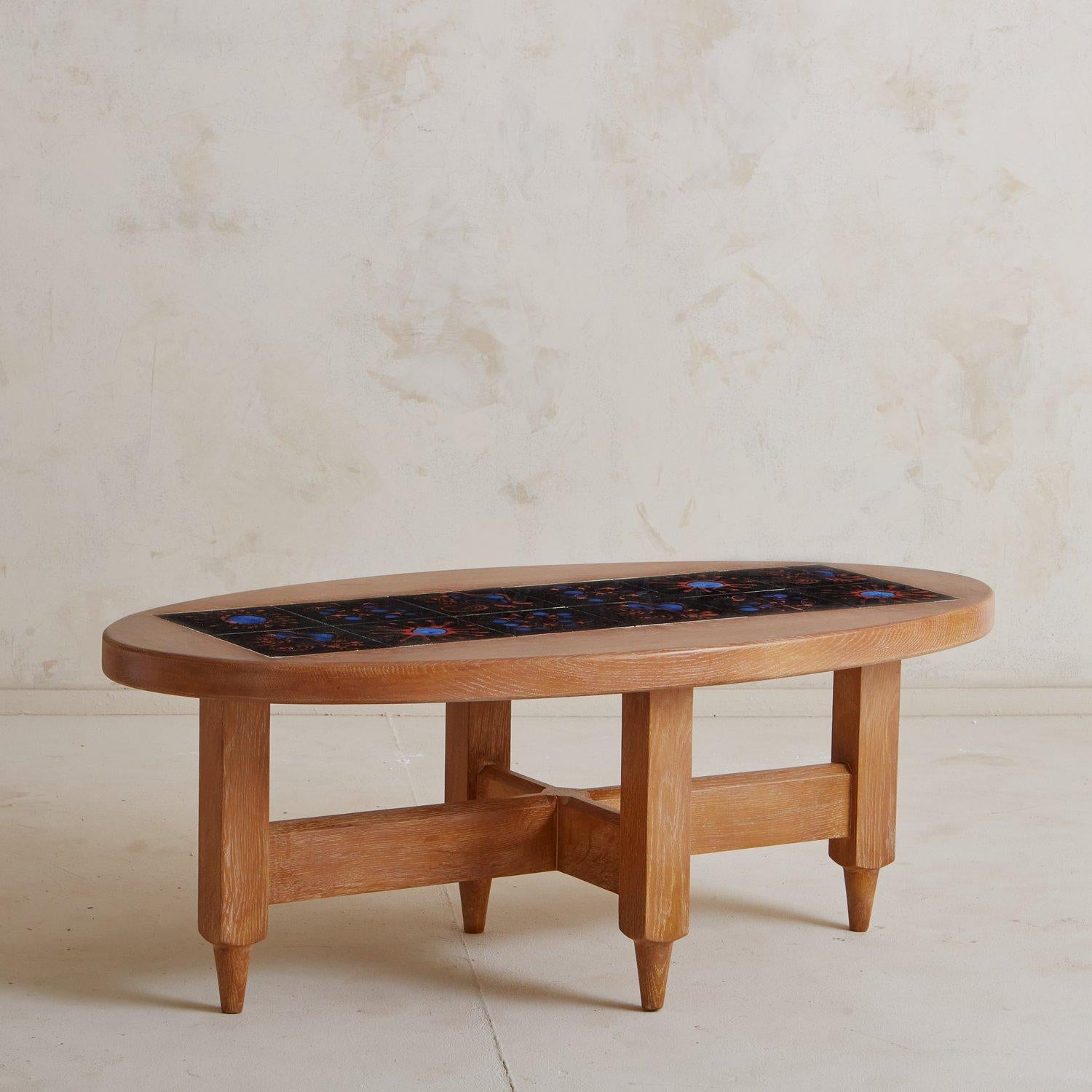 A Mid Century French coffee table by Guillerme et Chambron constructed with oak. This timeless piece has an oval top with a black ceramic tile inlay featuring blue and red abstract patterns. We love the sculptural carved base and X-support.