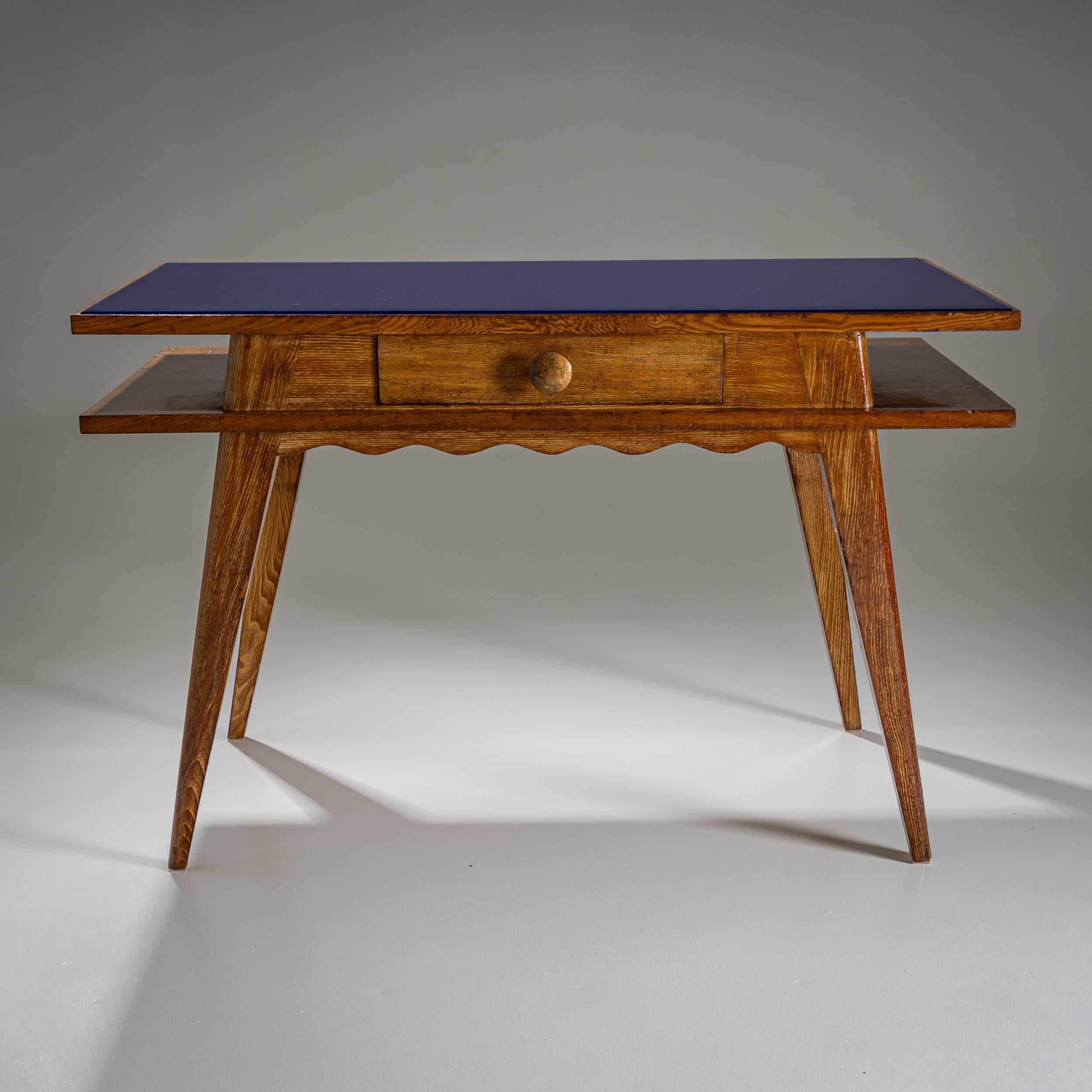 Escritorio de estilo de mediados de siglo con un elegante tablero de cristal azul y cuerpo de roble. Un cajón central con un pomo redondo de madera ofrece espacio de almacenamiento. Las patas abocinadas y cónicas y el delantal festoneado realzan el