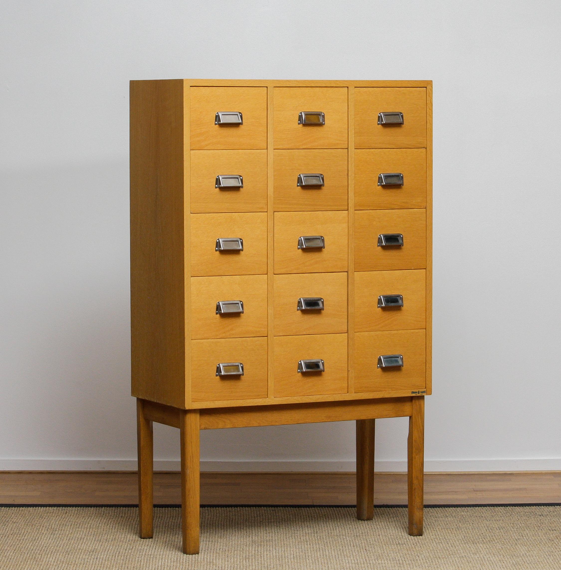 Dies ist ein Archivschrank aus Eichenholz, hergestellt von Lövgrens in Tarred, Schweden. Der Schrank hat ein modernes Mid-Century-Design mit fünfzehn kleinen Schubladen, die jeweils mit Label-Haltern und Griffen aus Metall ausgestattet sind. Seine
