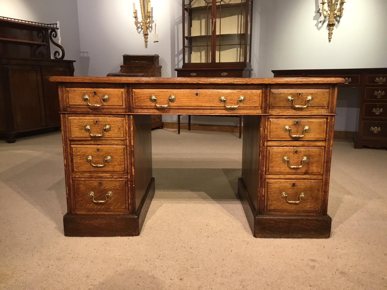 An oak Edwardian period antique pedestal desk. Having a rectangular solid oak top with a tan leather inset writing surface with blind and gilt tooled detail, above nine rectangular mahogany lined drawers each having traditional brass swan neck