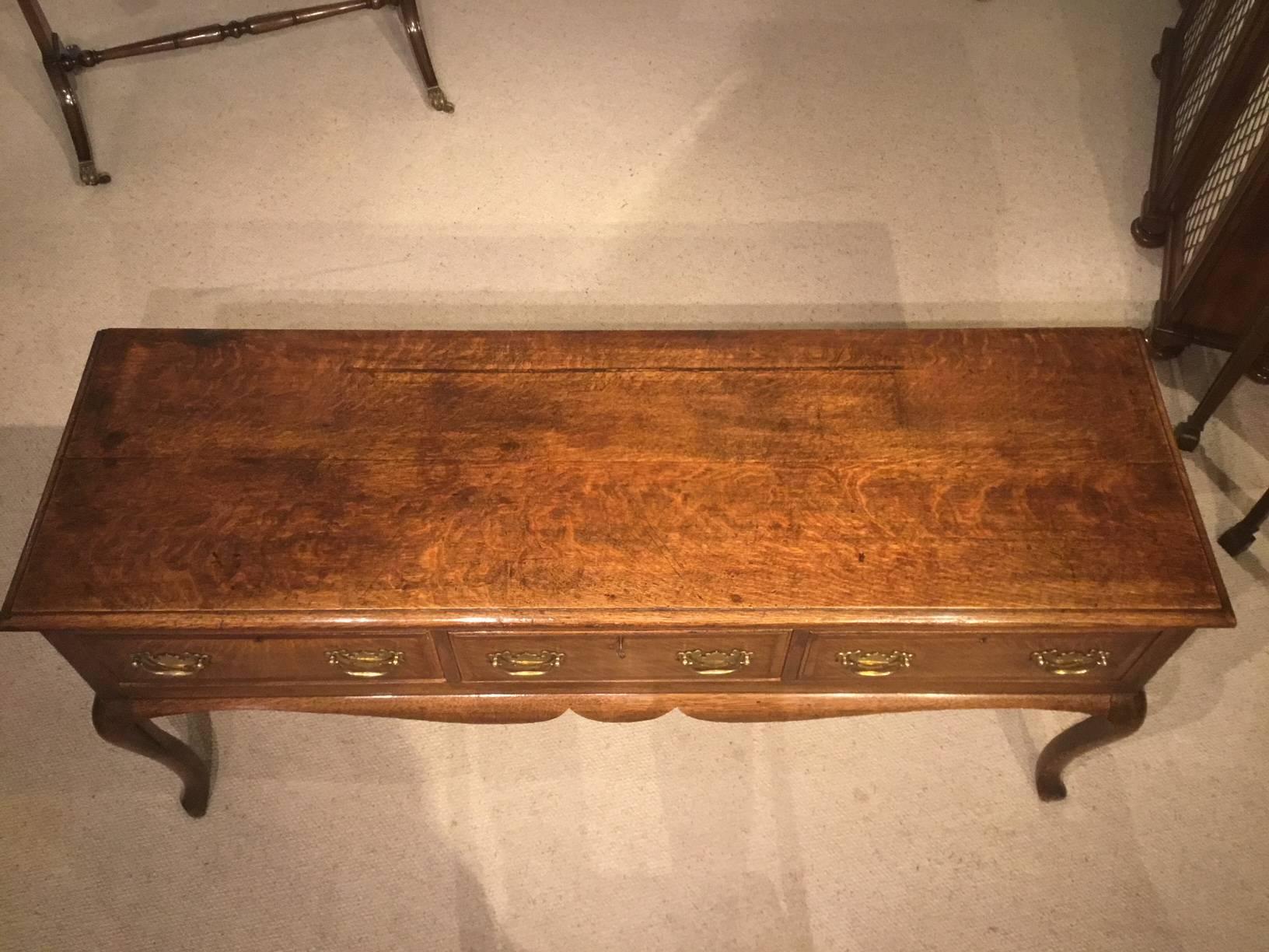 An oak George III period antique dresser base. The rectangular two plank top above an arrangement of three oak lined drawers with mahogany crossbanding to the oak fronts and traditional brass swan neck handles, with a wavy frieze the whole supported