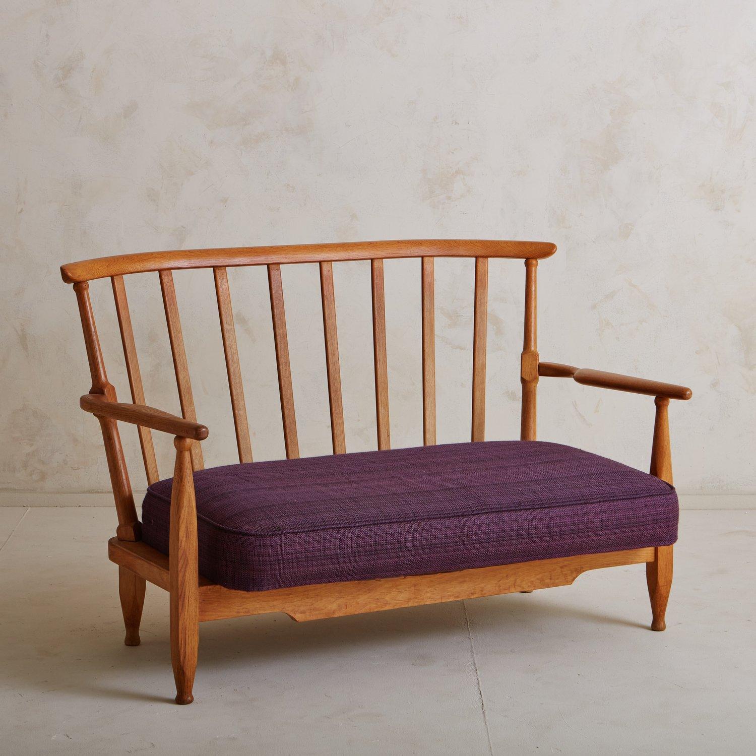 A Mid Century French settee by Guillerme et Chambron constructed with oak. This timeless piece features beautiful carved details and an elegant, angled spindle back. It has two removable cushions in their original purple fabric. Unmarked. Sourced in