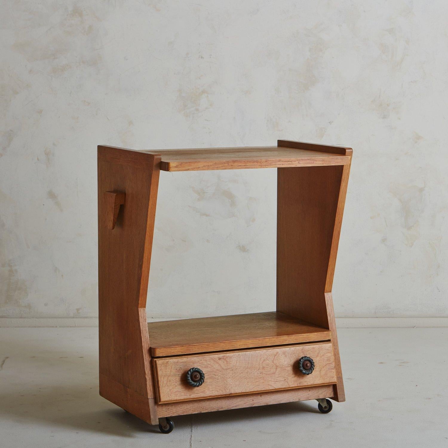 A sculptural 1960s oak side table by French design duo Guillerme et Chambron. This table has an angular profile with an open center and one bottom drawer. It has beautiful braided metal hardware and stands on casters for easy mobility. Unmarked.