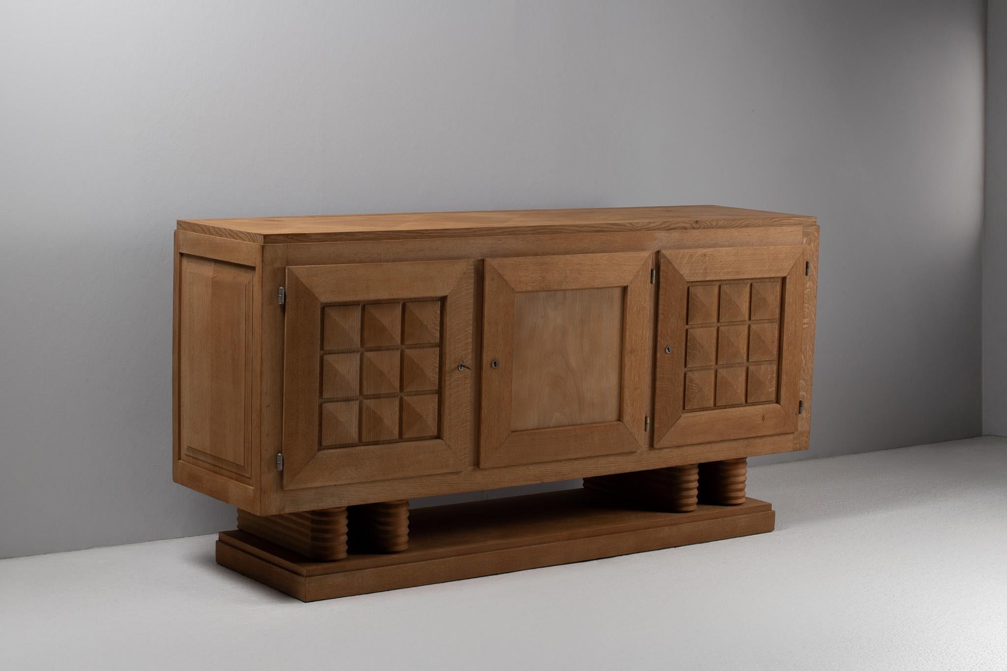 Sculpted in stoic relief yet softened by time, this oak sideboard by Gaston Poisson asserts a quiet monumentality. Its architectural presence is anchored by fluted cylindrical feet, while the front façade alternates between stark paneling and