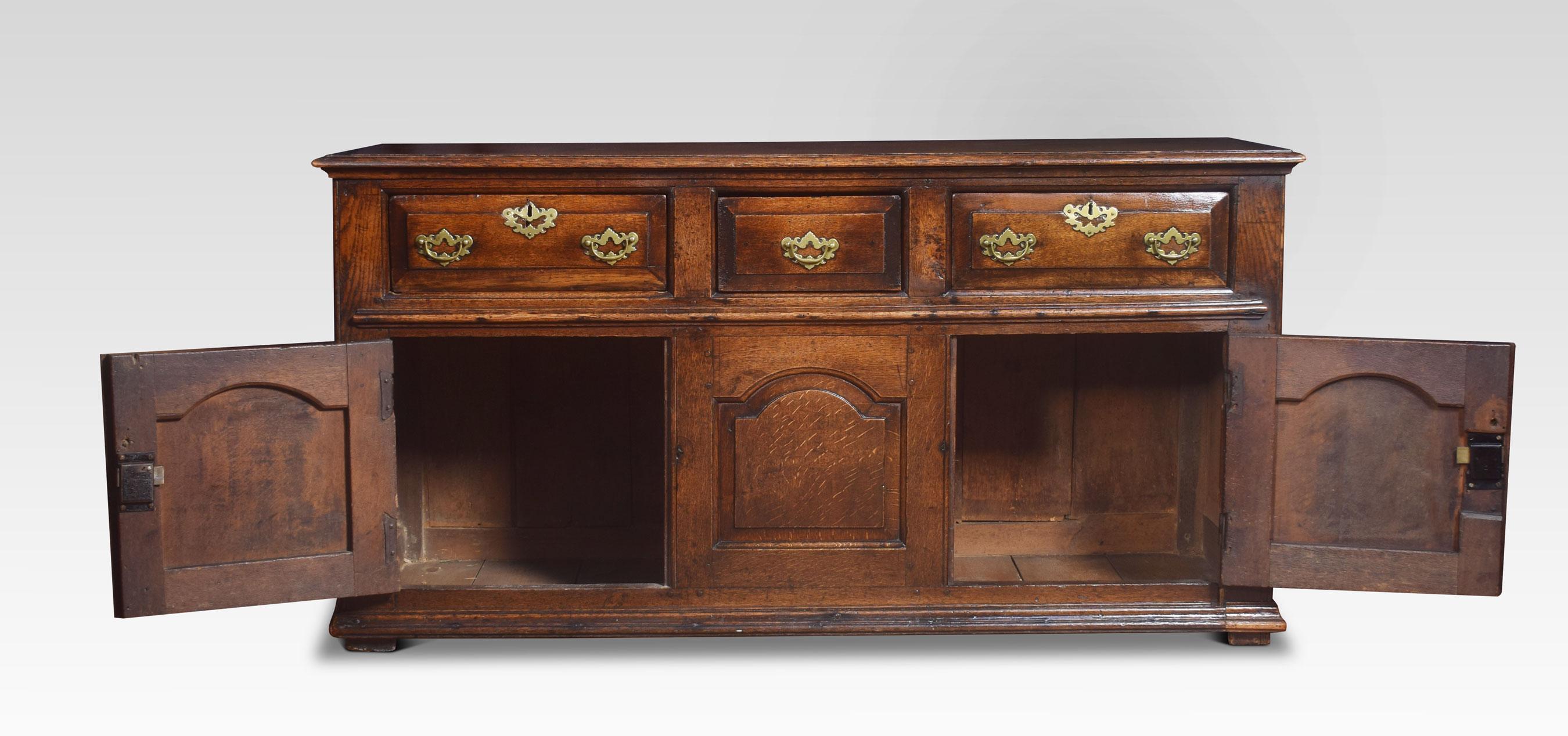Oak dresser base, the large rectangular top having quadrant mould edge. To the frieze fitted with three frieze drawers each with chamfered edges and brass drop handles and pierced backplates. Above two cupboard doors with framed arched paneled doors