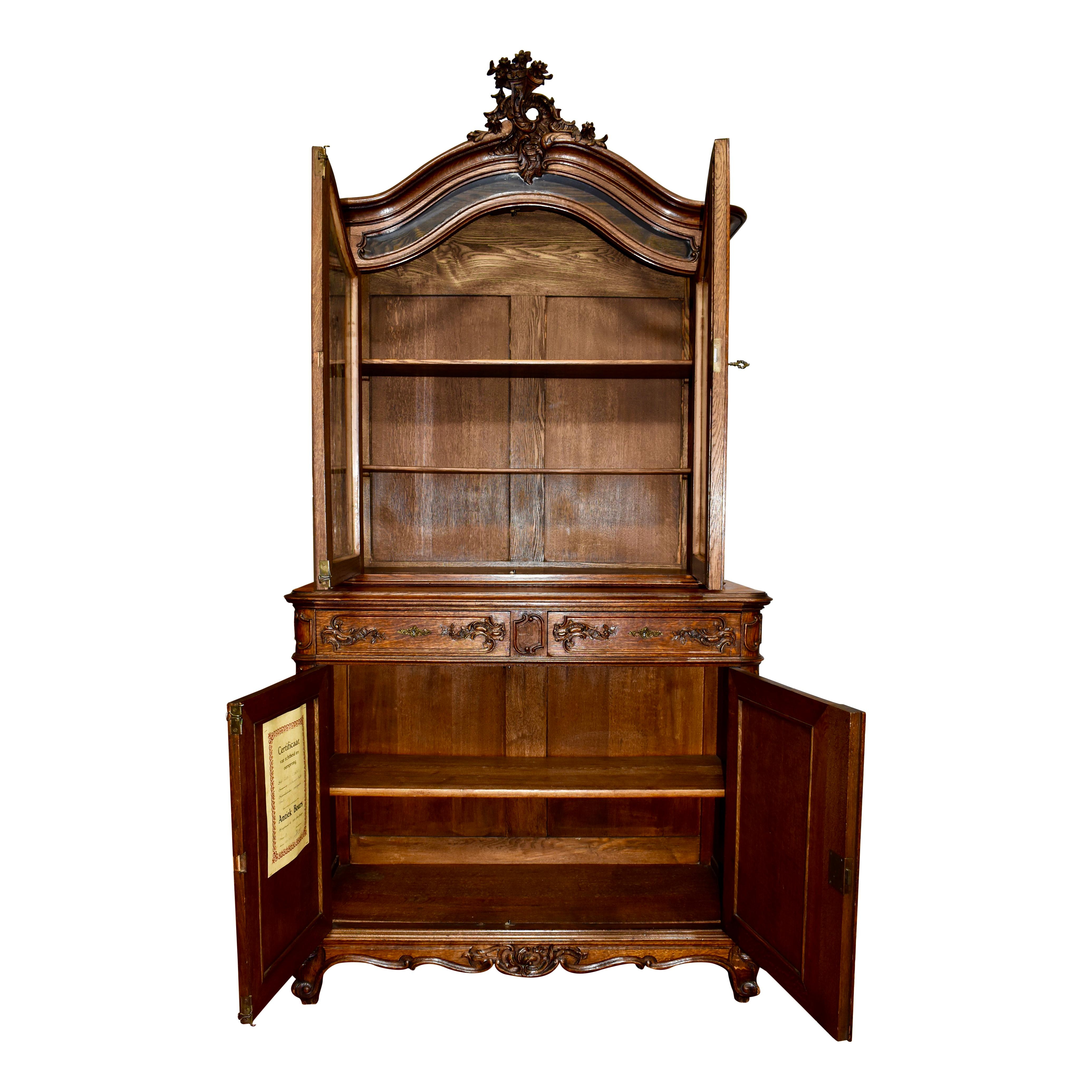 An arched cornice culminates in a beautifully pierced, foliated design incorporating flowers, ruffles, and scrolled leaves atop this oak vitrine. The arched, double doors with glass panes of the upper gallery are overlain with pierced foliated