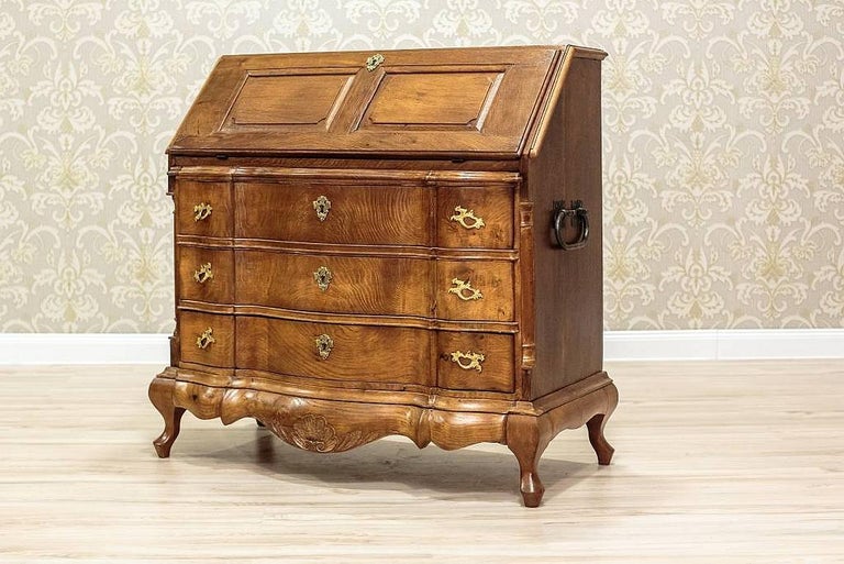 This neo-baroque writing desk is entirely made in oak wood. Presented piece of furniture is dated the end of the 19th century (circa 1890). The item is composed of two segments: a liftable upper part, a chest with a row of three drawers, and a fall