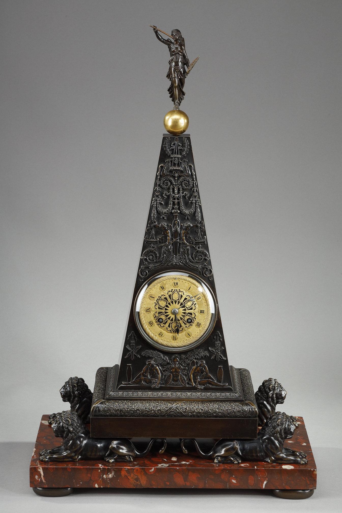 Very large clock, halfway between a pyramid and an obelisk, in patinated and gilded bronze, resting on four antique-style lions and a cherry-red marble veneer base. Bas-relief ornamentation of leafy garlands, trophies of arms, surmounted by a figure