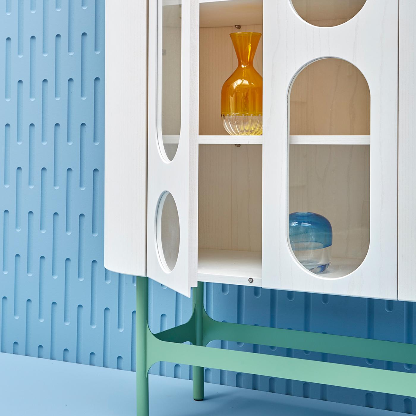 Boasting an elegant rounded silhouette, the Oblò B Cabinet designed by Matteo Zorzenoni has a white-lacquered ash wood frame raised on a four-legged metal base in a pastel blue finish. The distinctive feature of the collection is the cut-out design