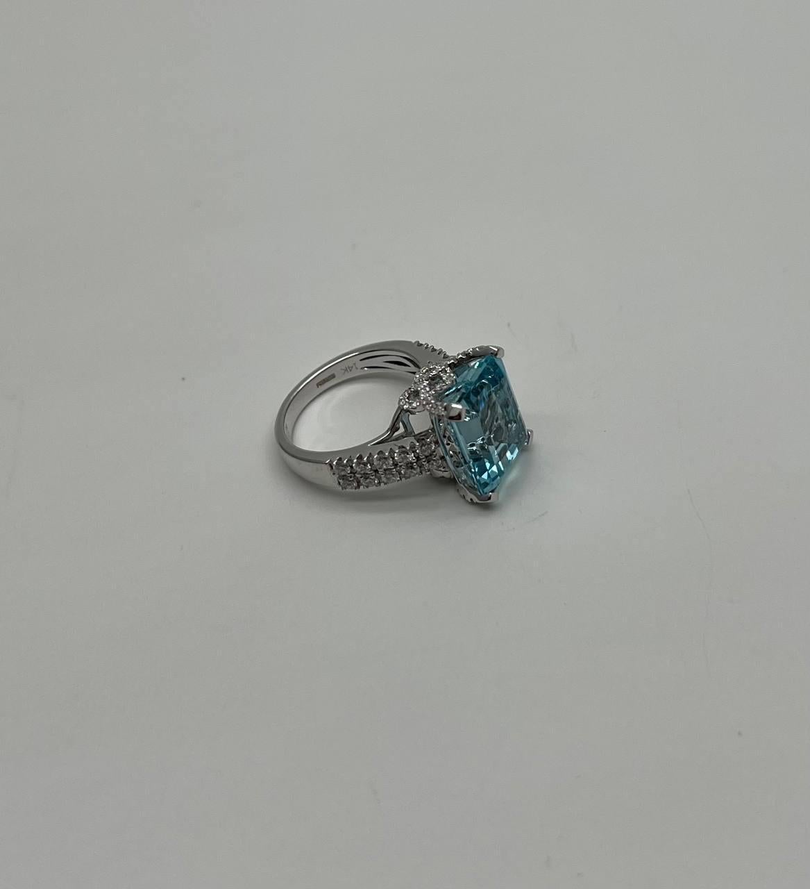 This elegant ring is crafted in the United States. It features a striking rectangular blue aquamarine, set in a polished metal band. The band is adorned with intricate detailing and smaller sparkling stones along the shoulders.
The gemstone is