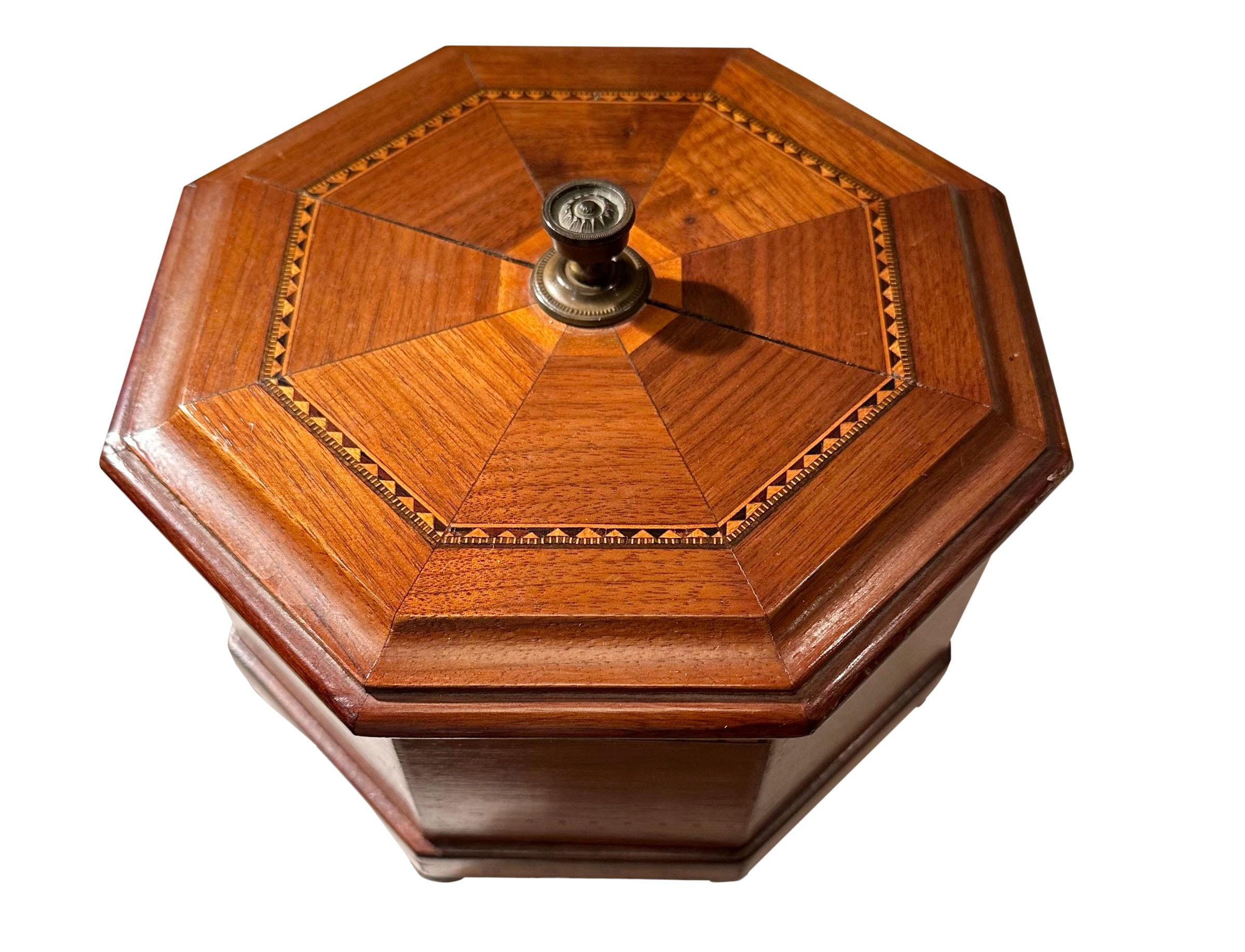 A lovely octagonal shape wooden box. The top with a striking inlay and a brass finial. Bun feet. The interior is three inches deep. English, turn of the century.