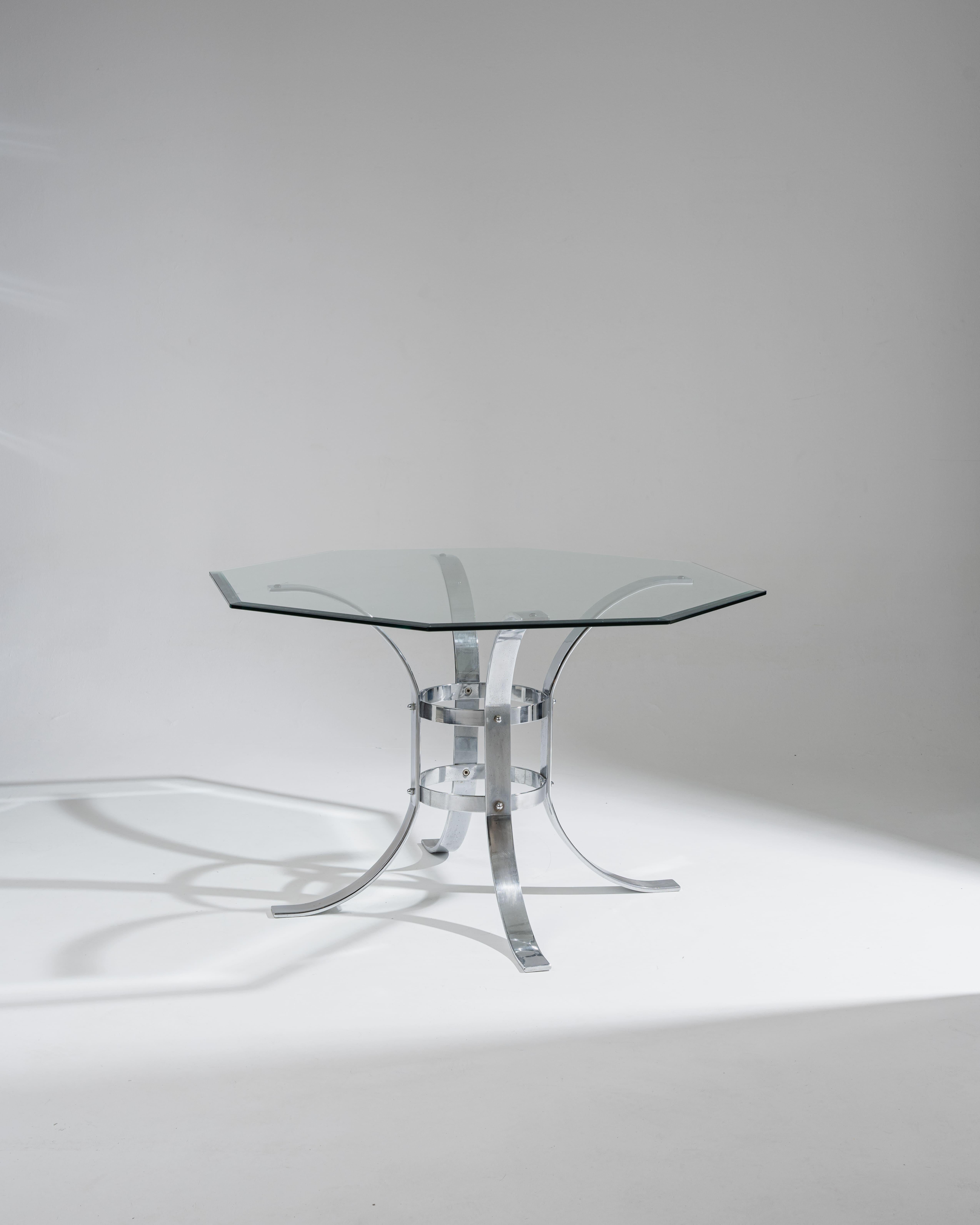 Mesa de comedor con tablero octogonal de cristal que descansa sobre una escultural base de metal cromado. La estructura presenta curvas dinámicas y travesaños centrales, típicos del diseño modernista de los años setenta. Muy buen estado general.