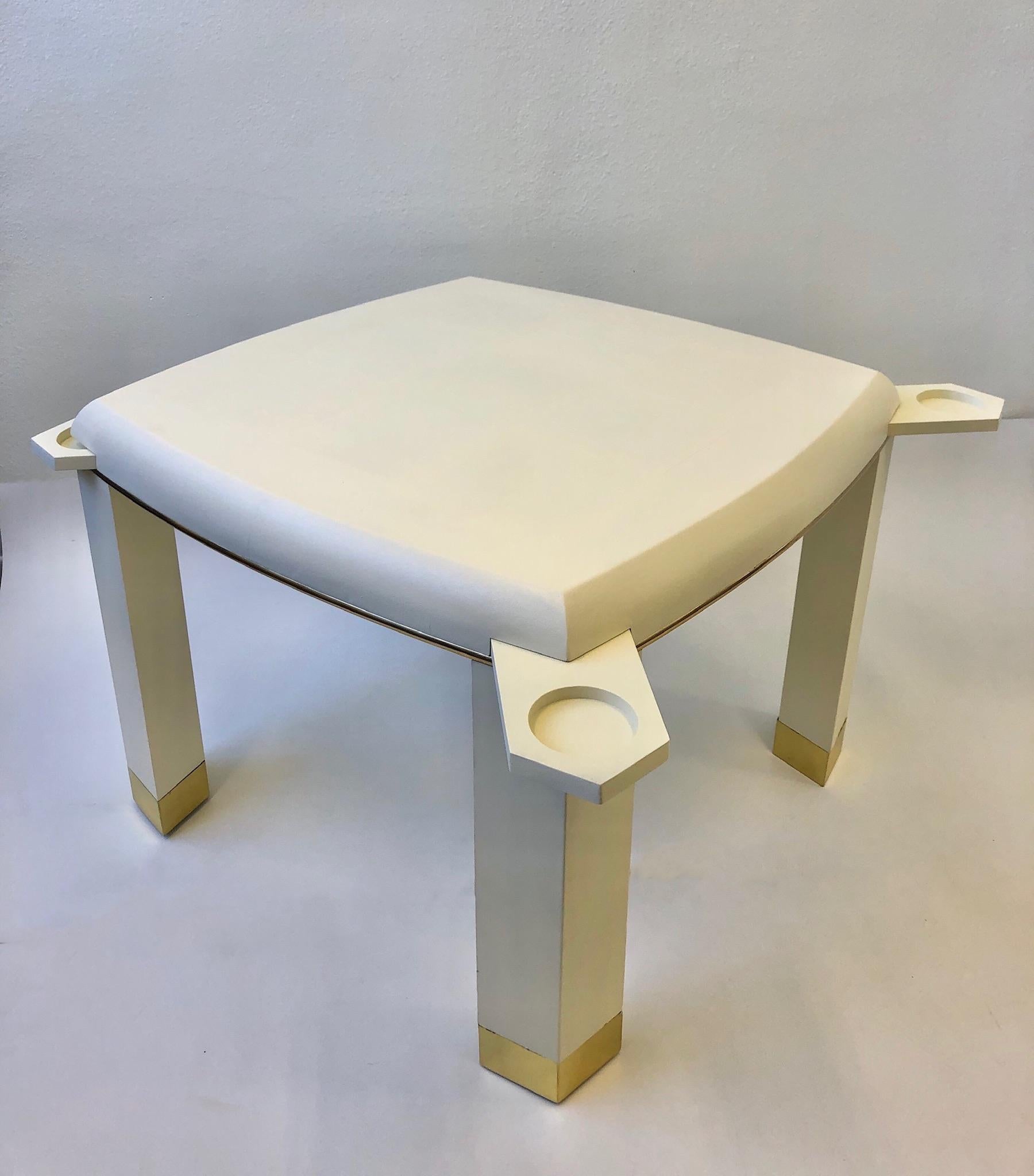 A glamorous game table with hidden drink or ashtray holder in the style of renowned designer Karl Springer in the 1980s. The table is constructed of wood that’s wrapped with off white snake embossed leather and polish brass detail. The table has a