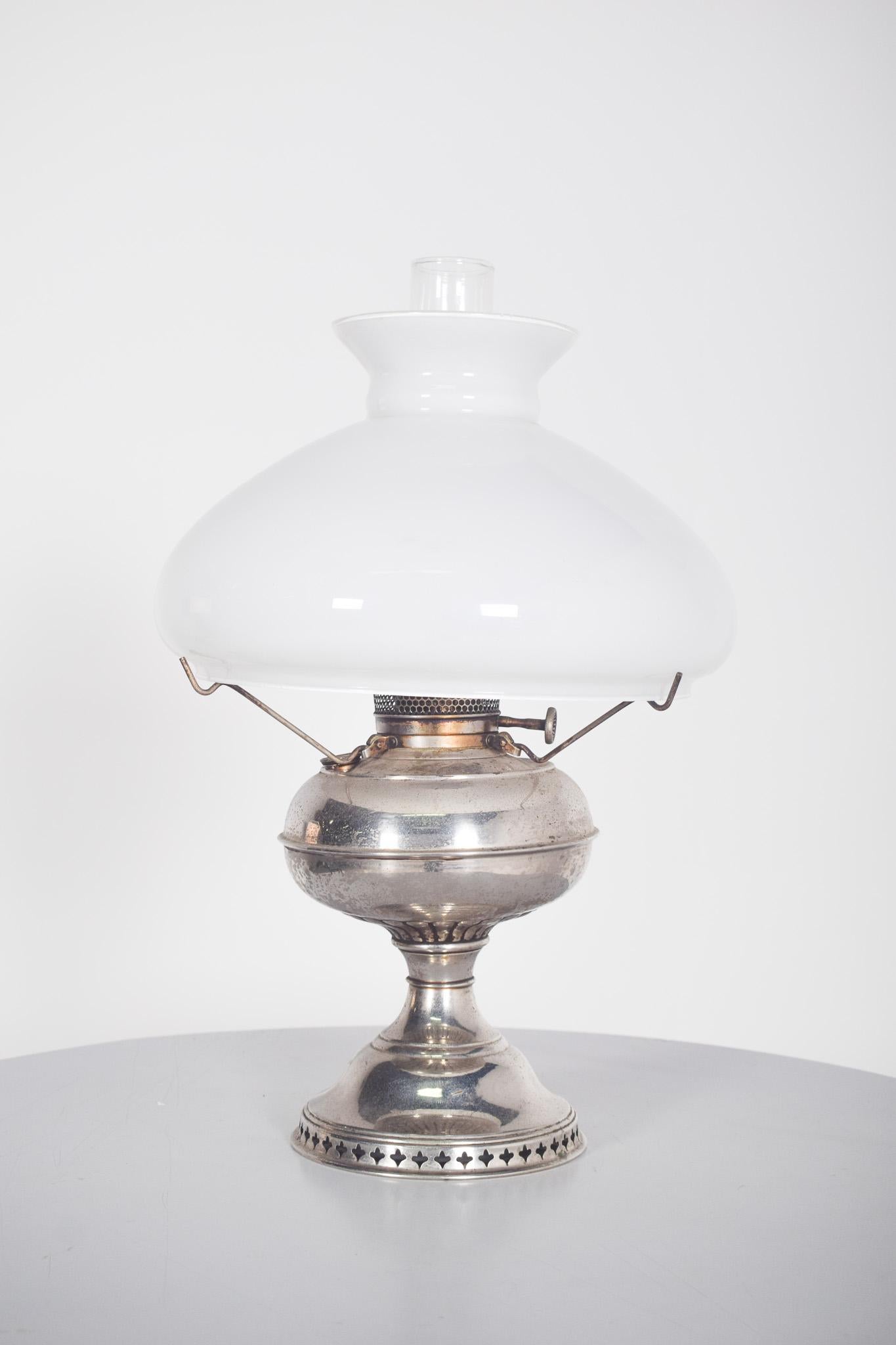 Lampe de table en métal chromé chauffée à l'huile. La base en métal argenté présente une finition vieillie, avec des détails perforés sur le fond, apportant une touche décorative et un charme vintage. Le corps arrondi soutient le mécanisme de