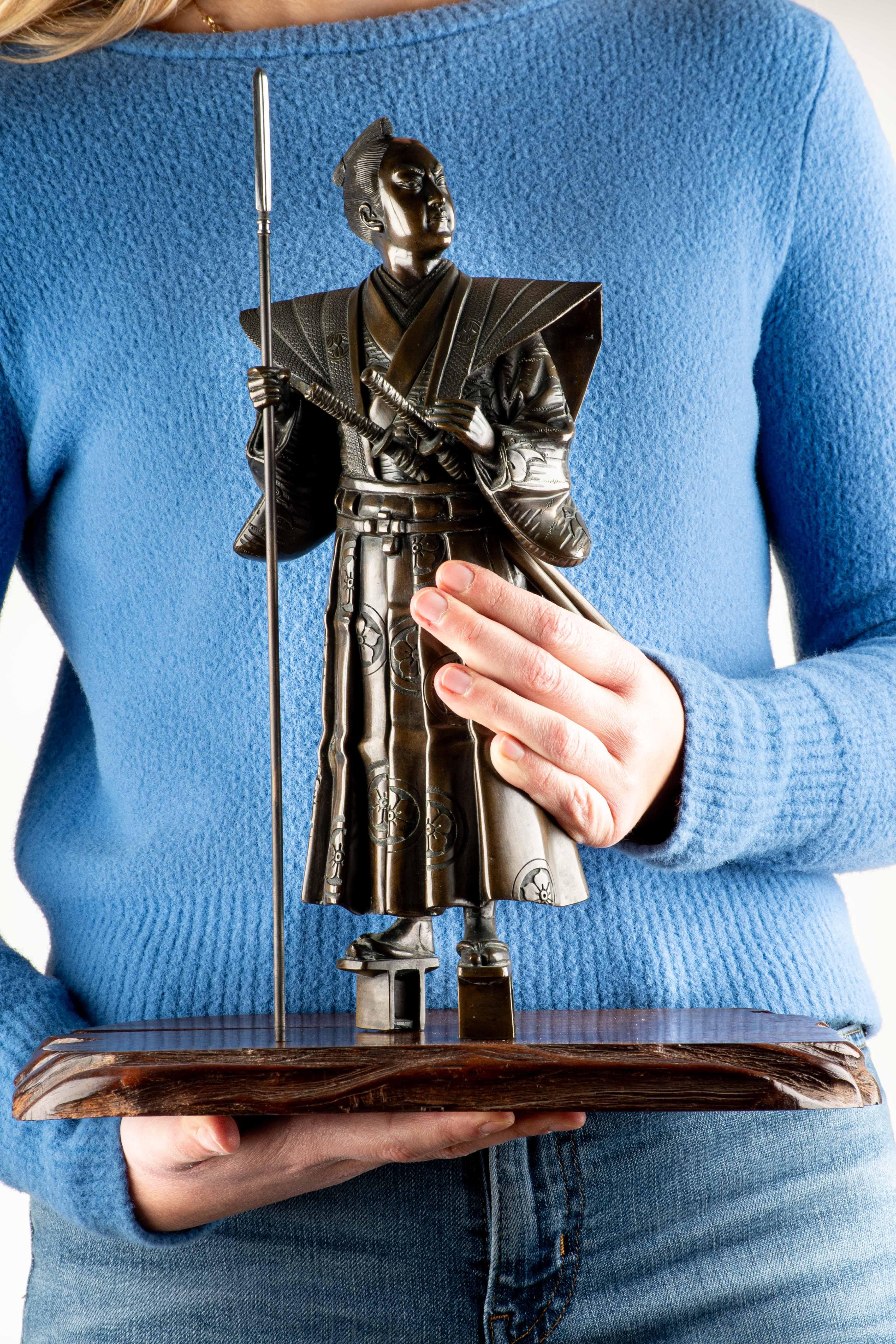 Imposing bronze sculpture depicting a majestic and solemn samurai in a standing position, holding a yari in his right hand. 

The samurai is dressed in a long kimono that wraps the body in flowing, elegant lines, highlighting the two swords: the