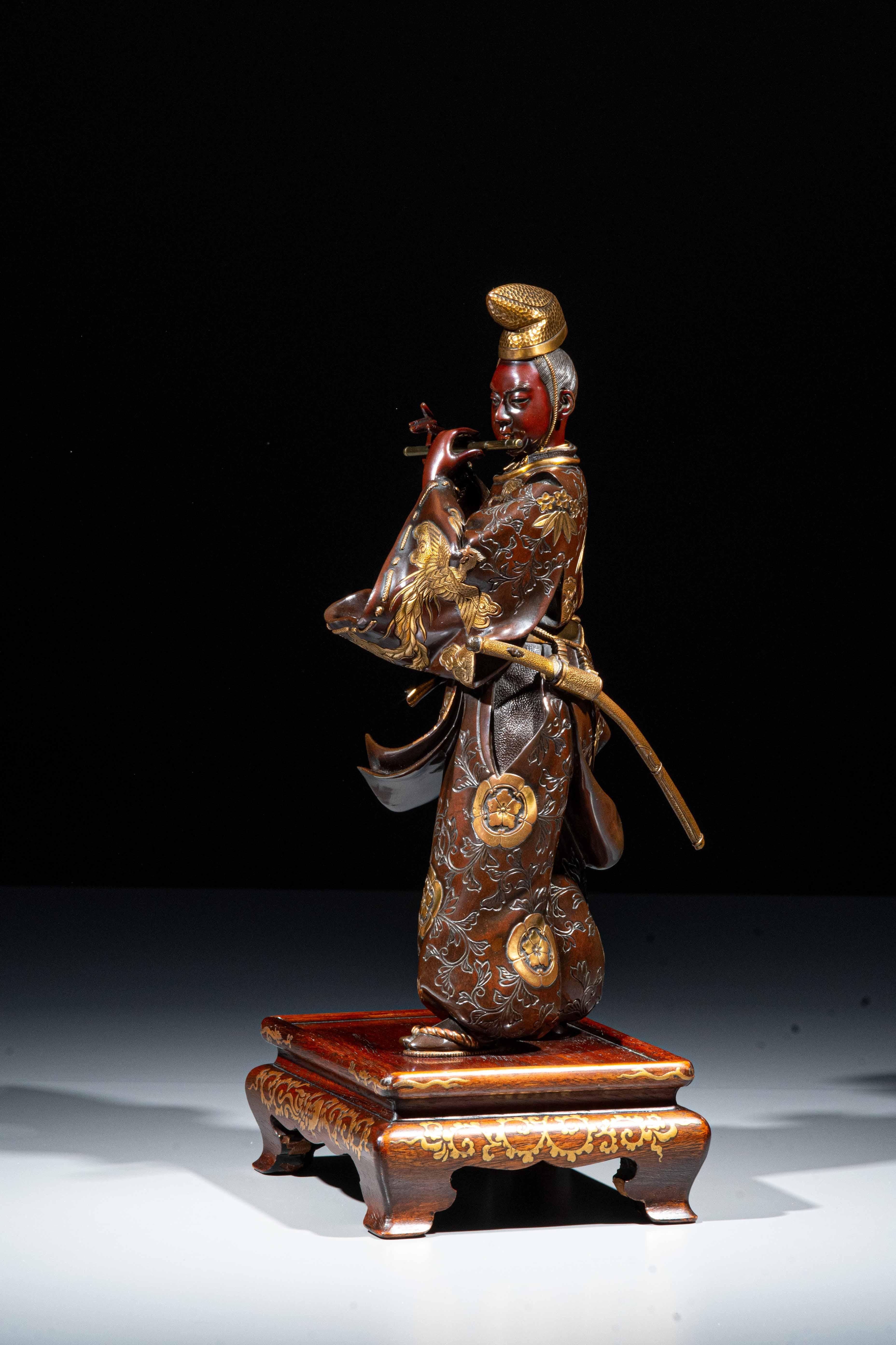 Okimono in bronzo patinato e dorato raffigurante il celebre guerriero Fujiwara no Yasumasa che suona il flauto, firmato Miyao. L’hitatare, mosso dal vento, mostra motivi di fenici e foglie firmato Miyao. La base in legno, classica delle opere di