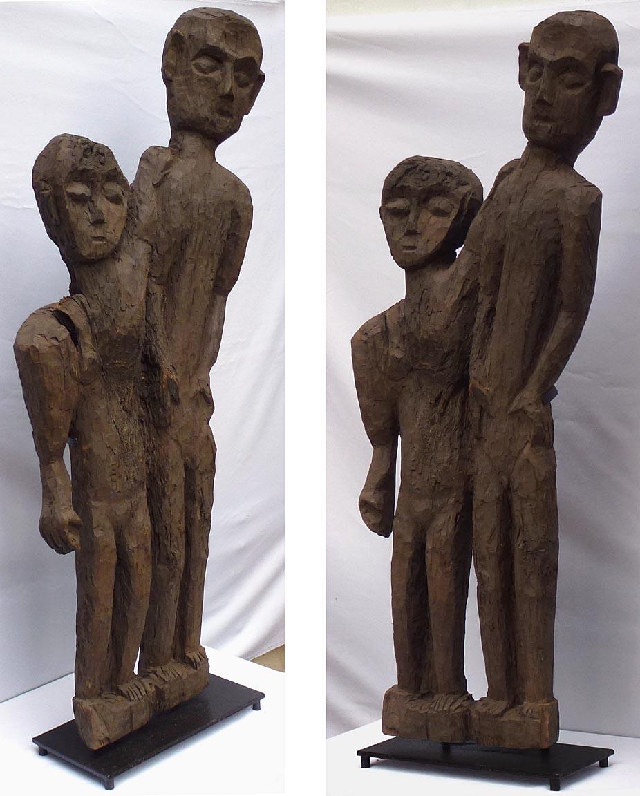 This is a 4 foot high naive and stylized carving of two people. It is roughly but expressively carved from a single slab of wood, mounted on a custom made metal base. It has a dark finish, probably paint. A knot hole in the wood passes through the