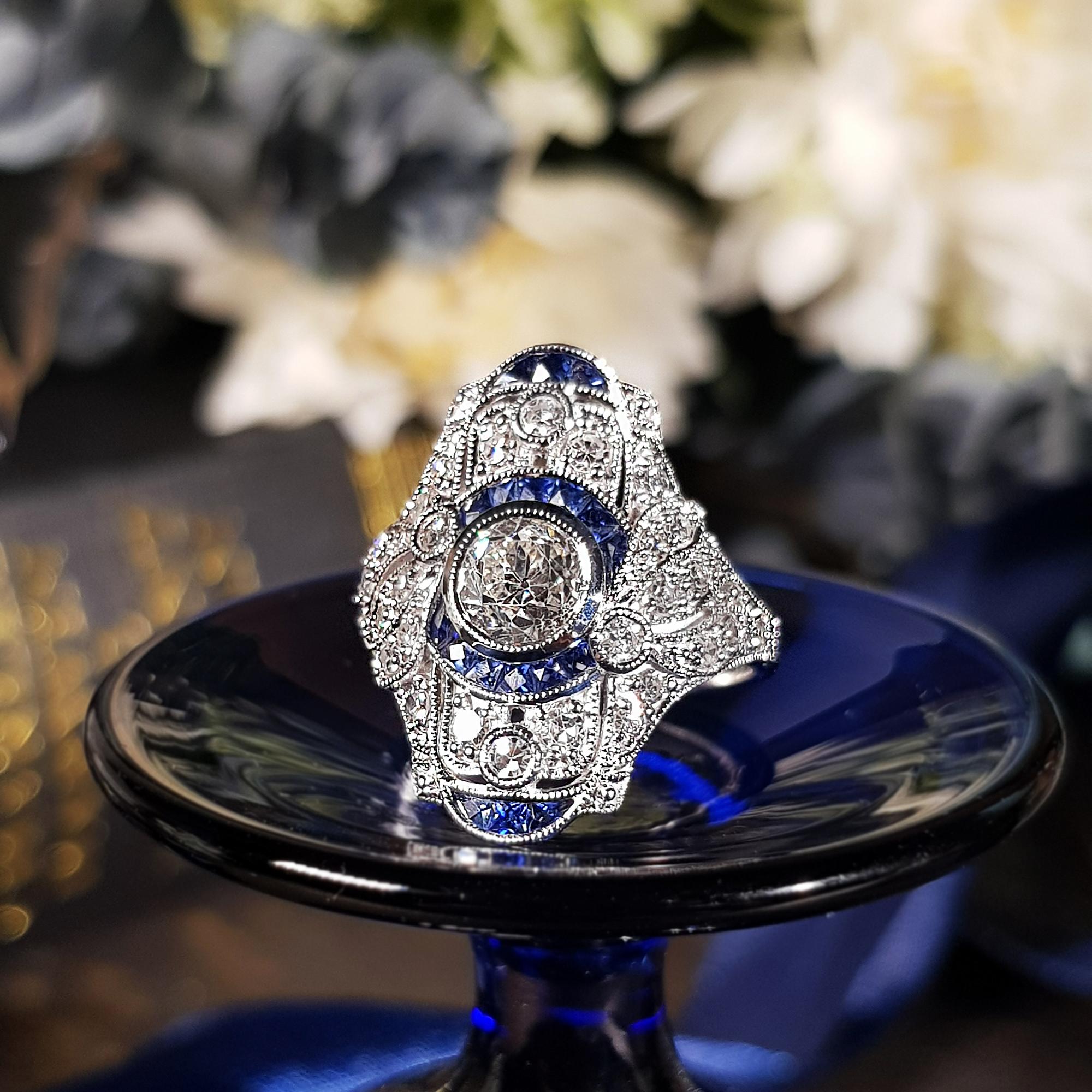 A strikingly stunning Art Deco design dinner ring sparkles the center with .99 old cut diamond accented the center with a round border composed of French cut blue sapphires. All are surrounded by single cut diamonds and more French cut sapphire on