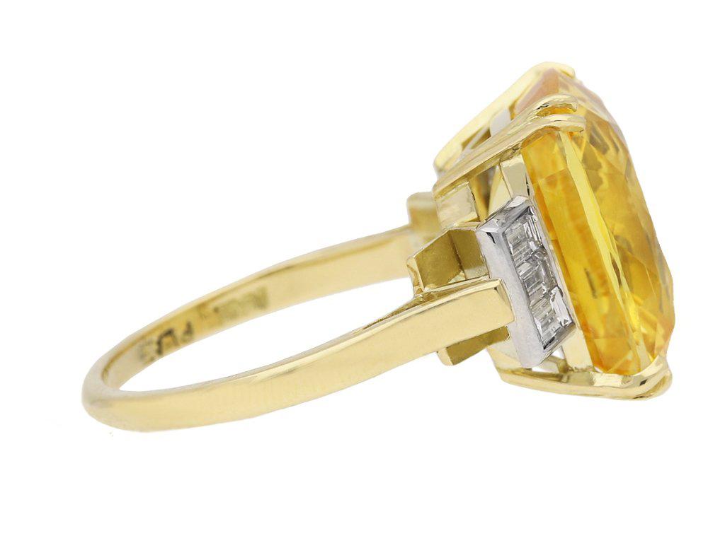 Yellow sapphire and diamond ring. Set with a cushion shape old cut natural unenhanced Ceylon sapphire in an open back four split claw setting with an approximate weight of 12.23 carats, flanked by six rectangular baguette cut diamonds in open back