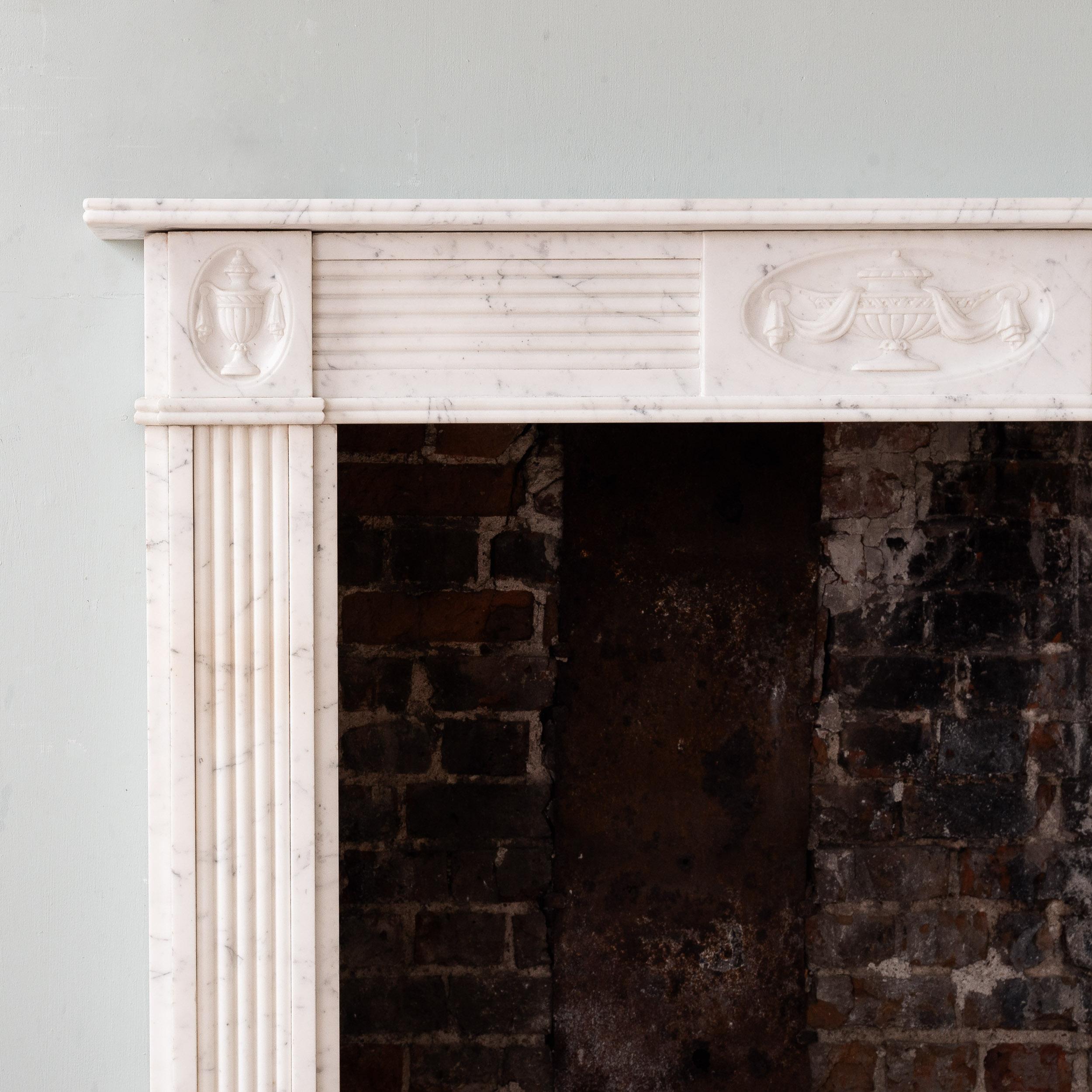 An ‘Old English’ Regency style fireplace, 1920s, with elegant reeded mouldings throughout, the frieze centred by relief carved tablet depicting a tazza draped in fabric.

Dimensions:	102.5cm (40¼