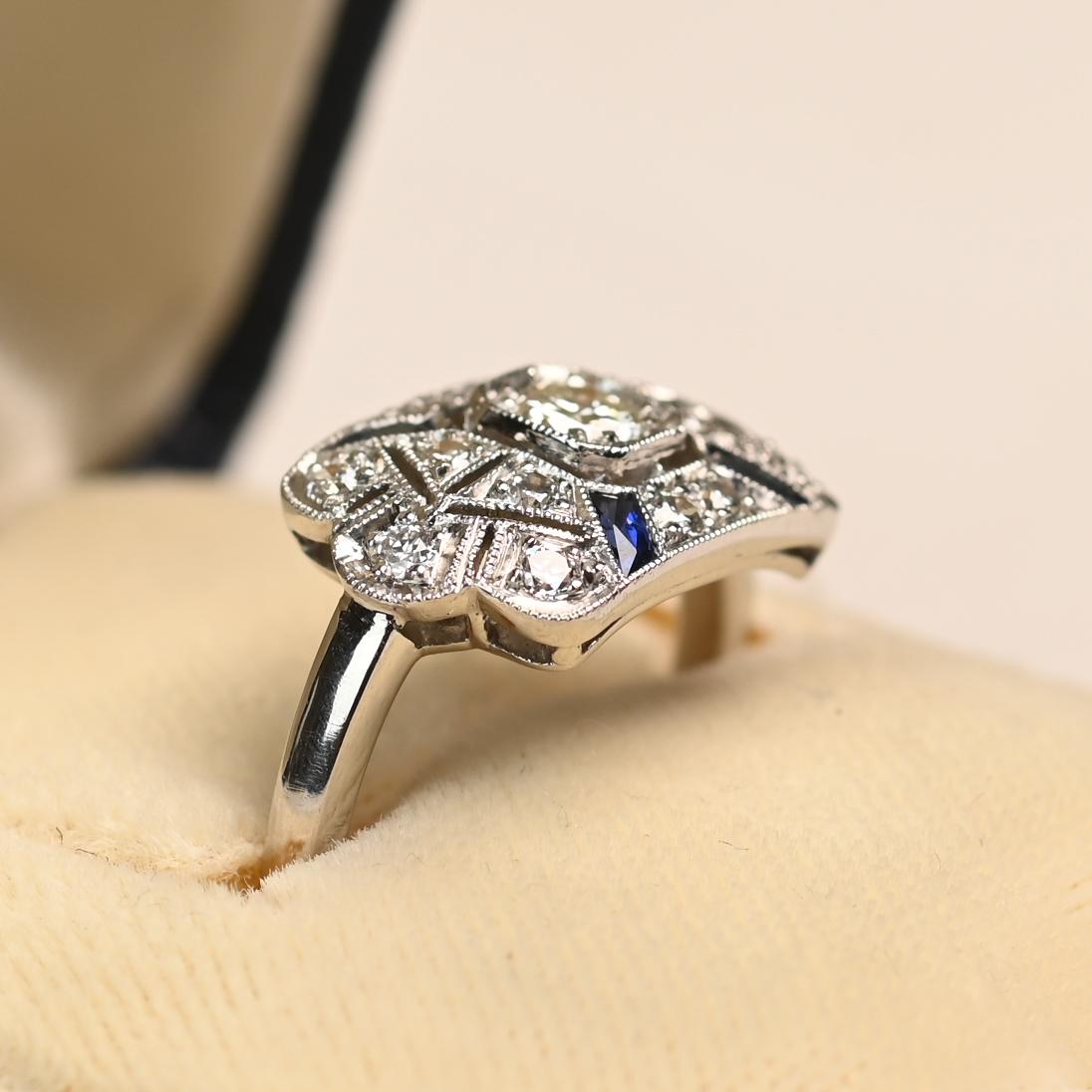 This exquisite Art Deco style ring centers on a lively old European cut diamond weighing approximately 0.20 carats, with J color and VS1 clarity. The central diamond is framed within an intricate geometric filigree design, beautifully accented by