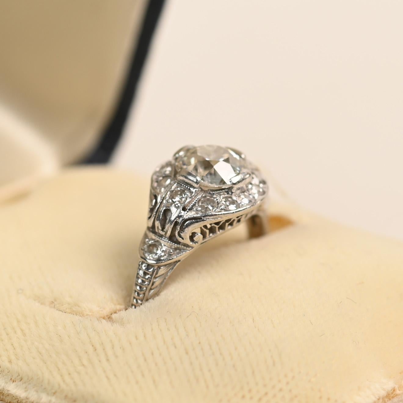 This Art Deco ring showcases a striking old European cut diamond at its center, weighing approximately 0.7 carats and graded J color with SI1 clarity. The diamond is beautifully framed by a geometric halo design, a hallmark of the Art Deco era, that
