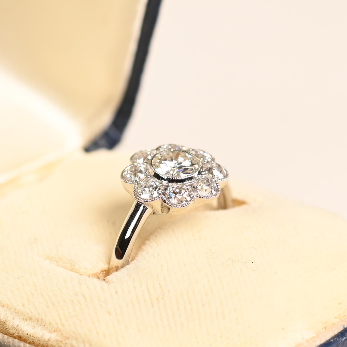 Este exquisito anillo de diamantes en racimo floral es una pieza artesanal atemporal, que muestra una armoniosa mezcla de encanto vintage y refinada elegancia. En su corazón yace un cautivador diamante de talla europea antigua, engastado en bisel