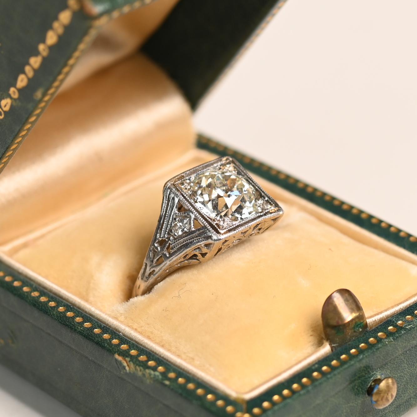 Este exquisito anillo Art Déco de platino, fabricado a mano hacia 1920, presenta un impresionante diamante de talla europea antigua de aproximadamente 1,50 quilates, elegantemente engastado en un bisel cuadrado. El diamante central, de color J y