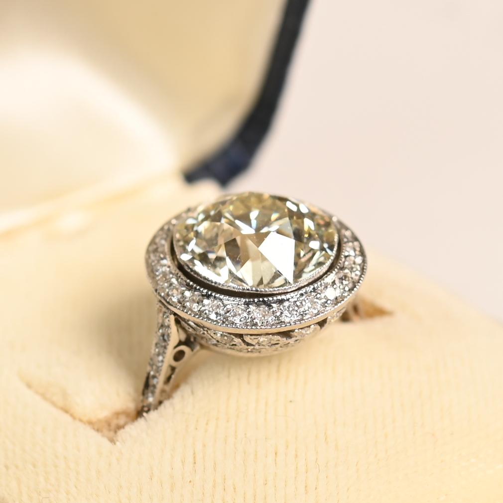 This exquisite engagement ring showcases a dazzling Approx. 4.30-Carats old European cut diamond at its center, graded L in color and VS2 in clarity. The antique-cut diamond is bezel-set and encircled by a graceful halo of pavé-set old European cut