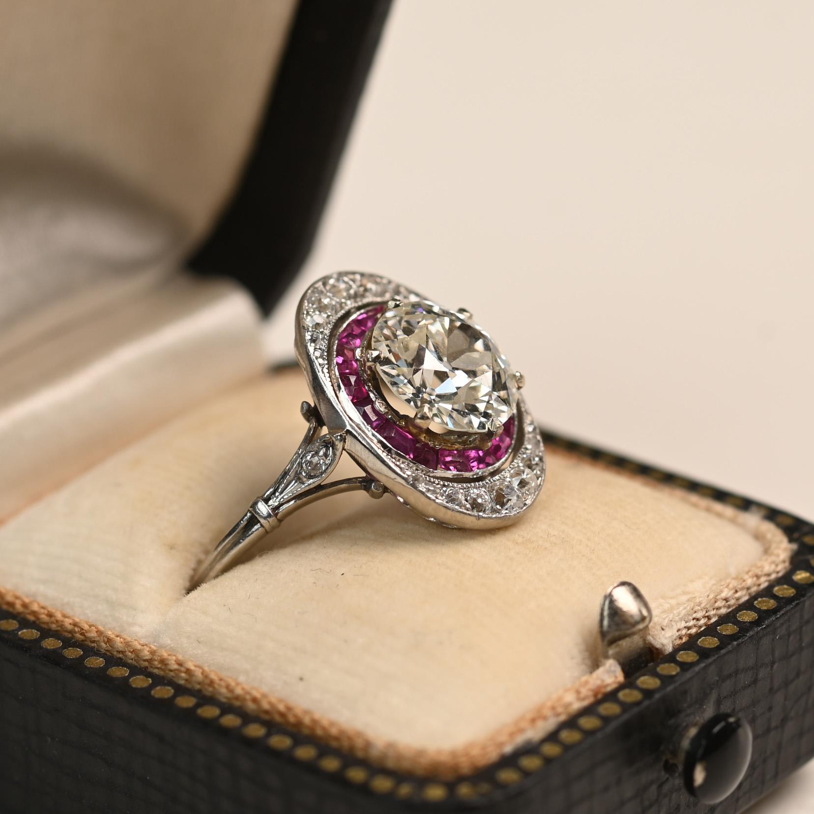 A striking Art Deco ring centering an approximately 2.10-carat Old European cut diamond, graded J color and VS1 clarity. The center stone is encircled by a halo of vivid French cut rubies, accented on the north and south points with Old European cut
