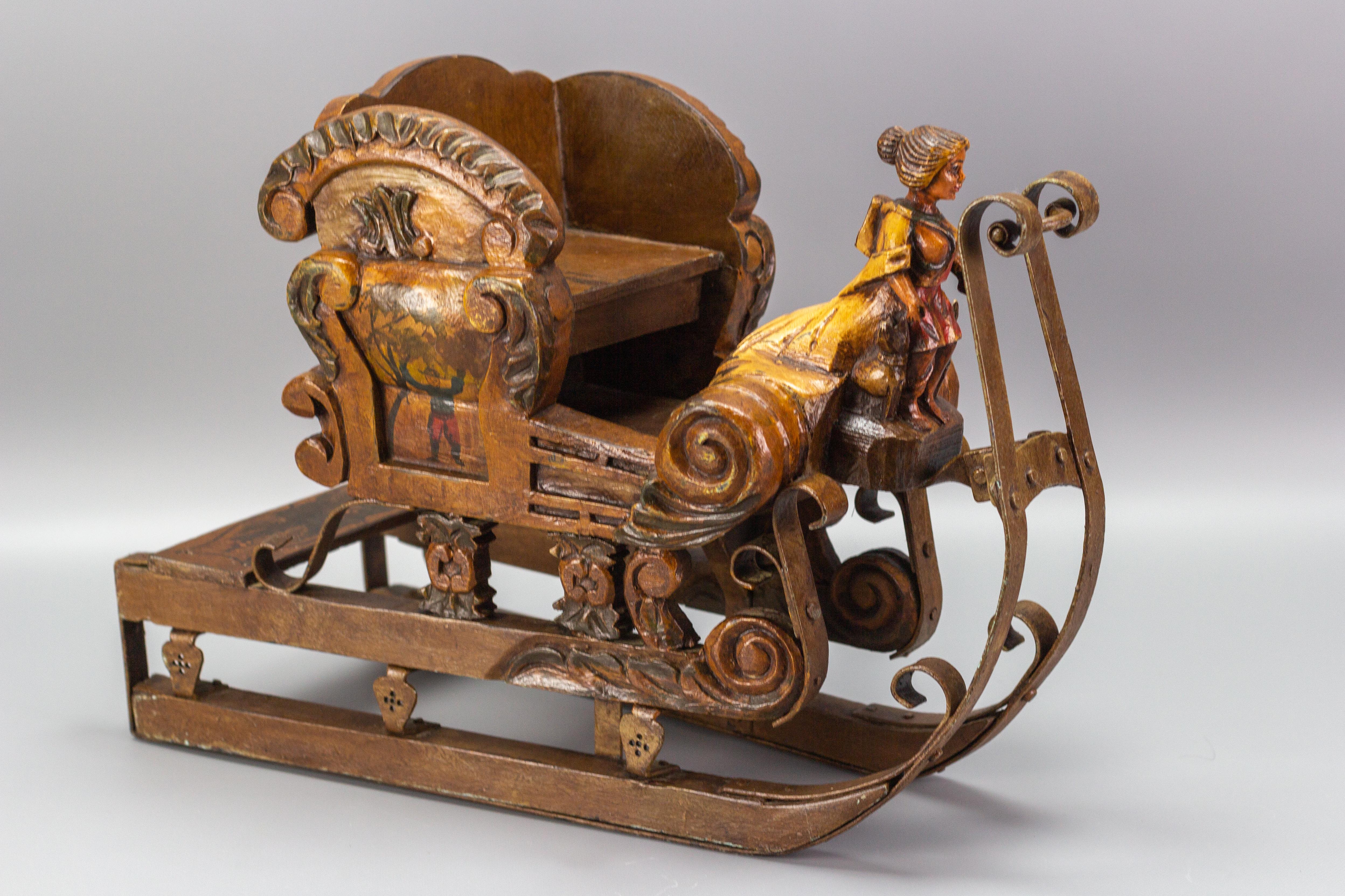 This gorgeous and handcrafted thick wooden sleigh features floral and foliate motif carvings and a carved woman figurehead at the front, flanked by two hounds; a seat for a doll behind, and a running board at the back. Iron runners over wooden with
